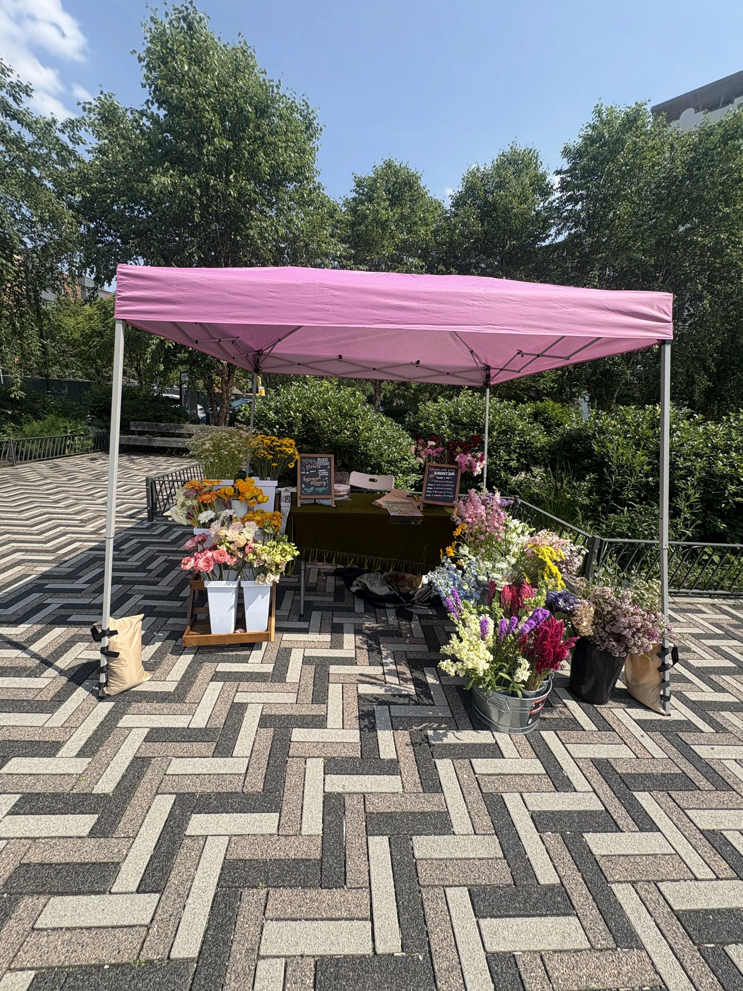 A flower stand under a pink canopy with various colorful flowers for sale in an outdoor park setting.