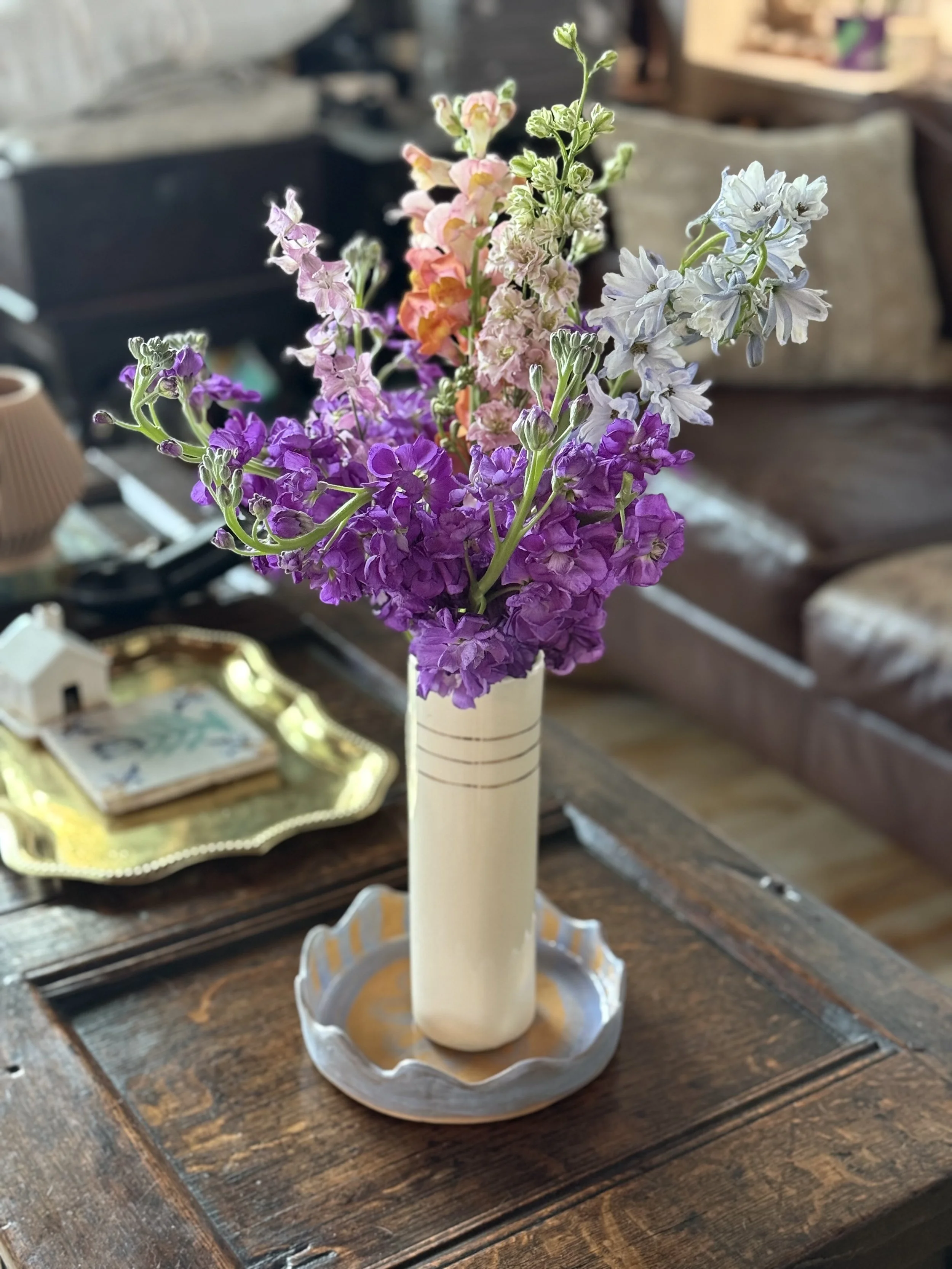 A white ceramic vase with vertical black lines contains a bouquet of purple, pink, yellow, and blue flowers, placed on a decorative tray on a wooden table.
