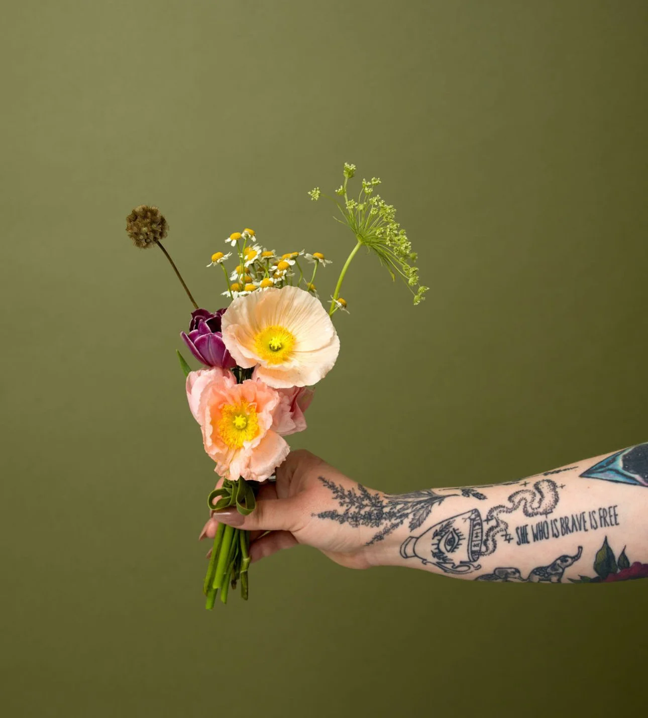 Hand holding a small bouquet of colorful flowers with tattoos on the arm, against a plain green background.