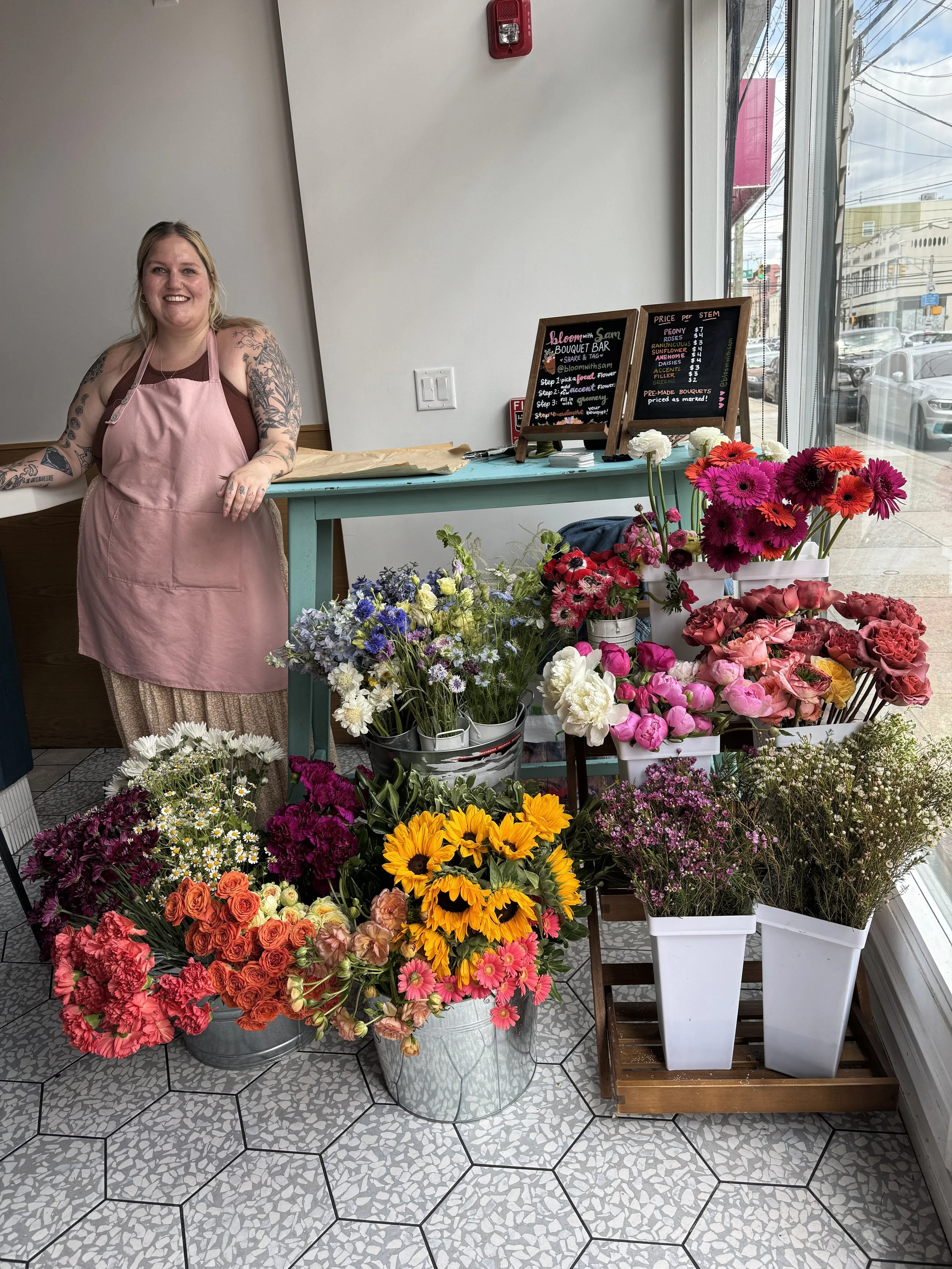 A smiling woman in a pink apron stands behind a display of colorful flowers inside a shop, with a street view visible through large windows.