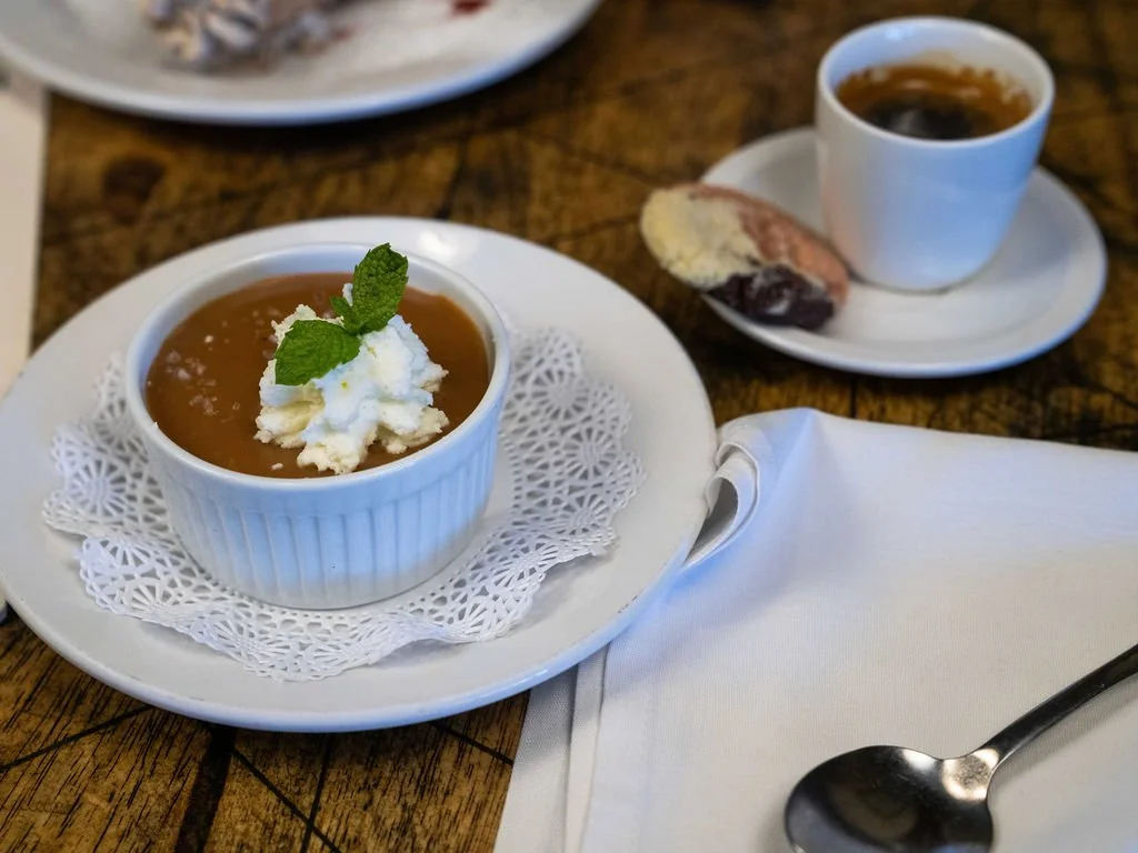 Butterscotch Budino topped with whipped cream and a mint leaf on a white plate with a doily, with a cup of espresso and cookies in the background on a wooden table.