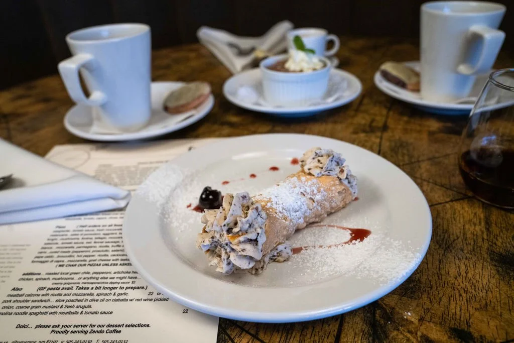 A dessert plate with a cannoli topped with powdered sugar, whipped cream, and a cherry, on a wooden table with coffee cups, a glass of red wine, and a dessert in a ramekin in the background