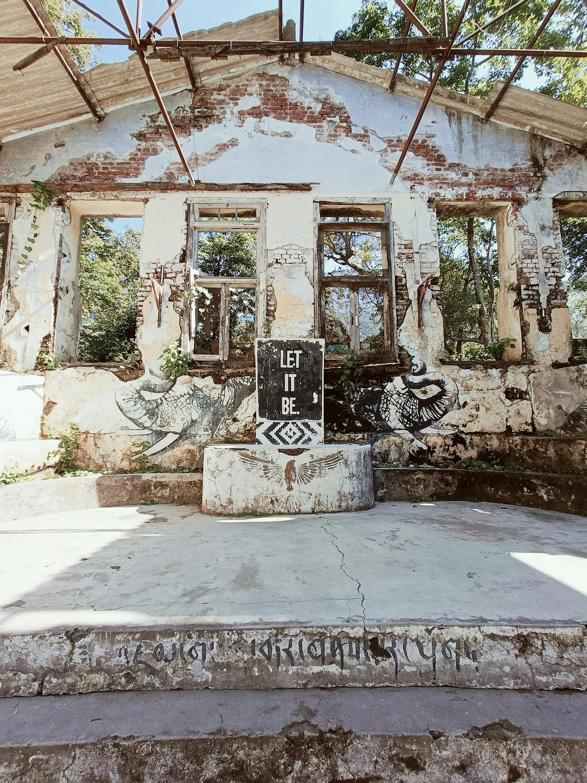 An abandoned, weathered building with peeling paint and broken windows, featuring graffiti art of elephants and an owl, a sign that says 'Let It Be,' and some handwritten script on the concrete steps.