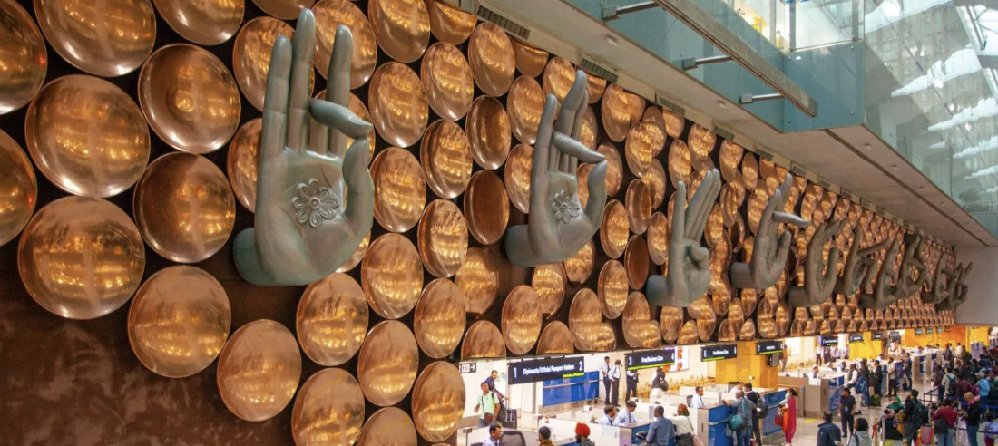 Decorative wall with large hands in different mudras placed among copper vases at an airport terminal with travelers at counters below.
