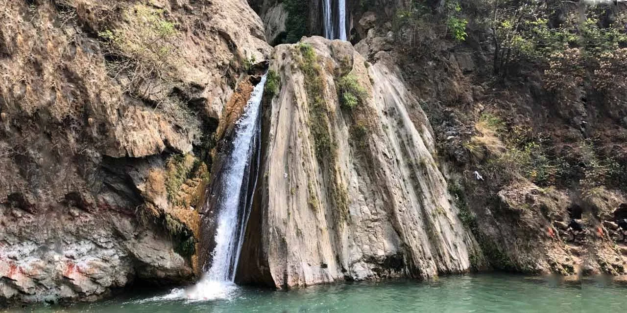 A small waterfall flowing over rocks into a green pool in a canyon with trees in the background.