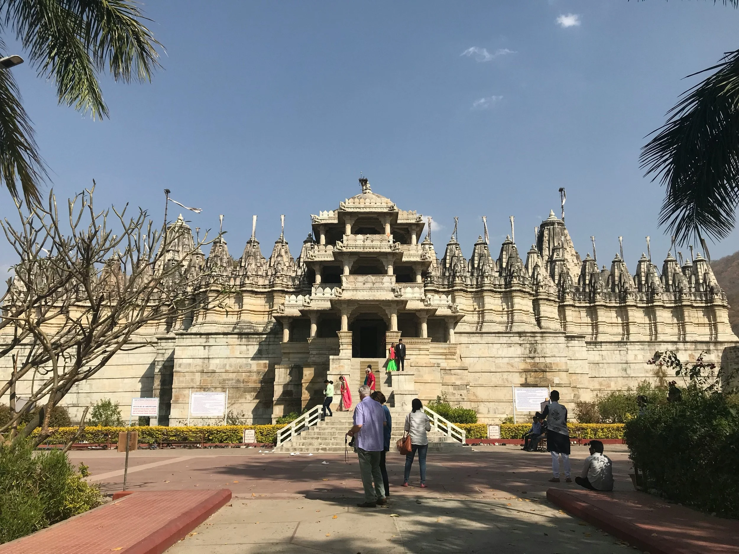 White marble temple with multiple spires, steps leading up to the entrance, and people walking around, with palm trees and a clear blue sky in the background.
