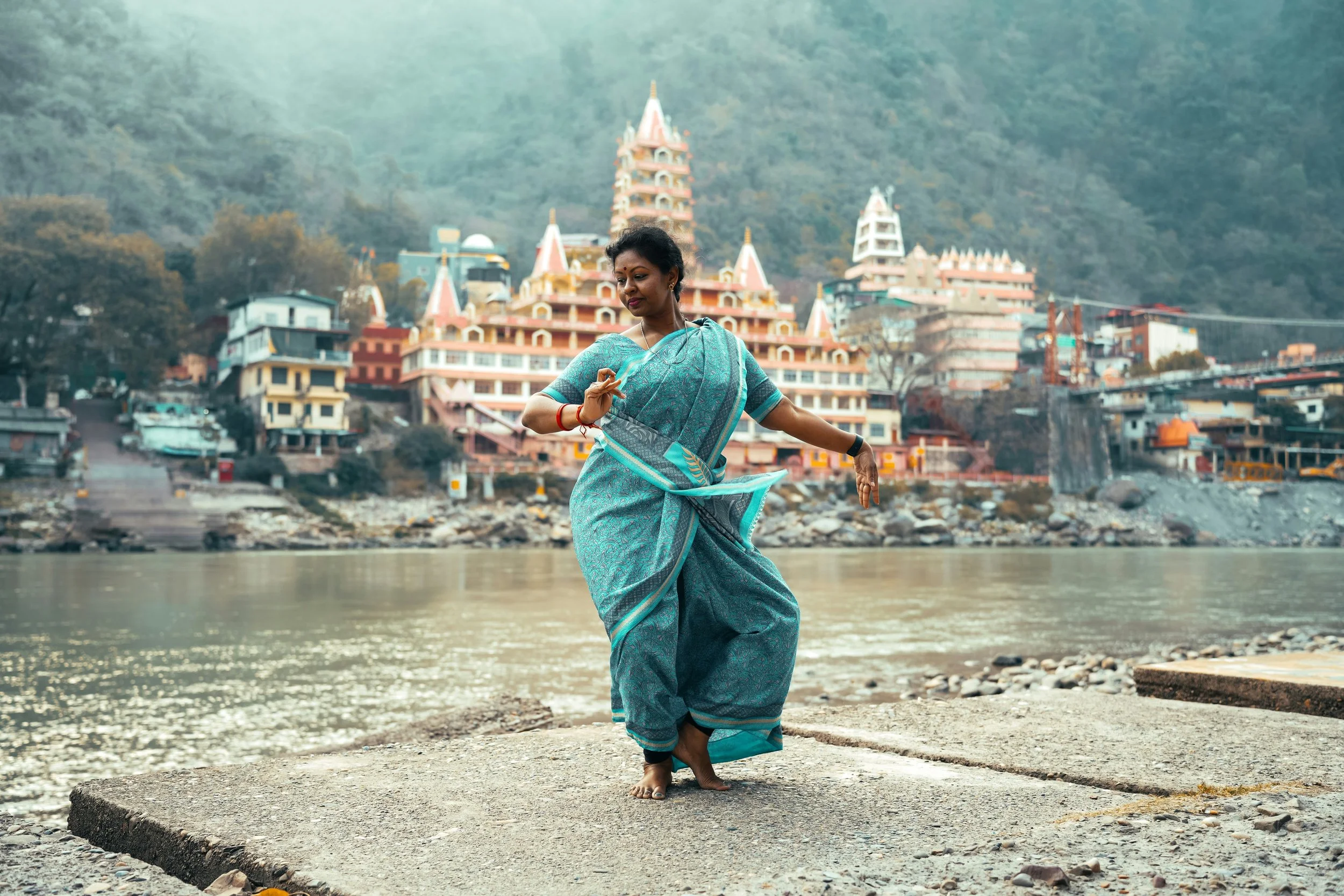A woman in a traditional blue sari dancing barefoot on a concrete platform by a river, with a large temple complex built into a hillside in the background.