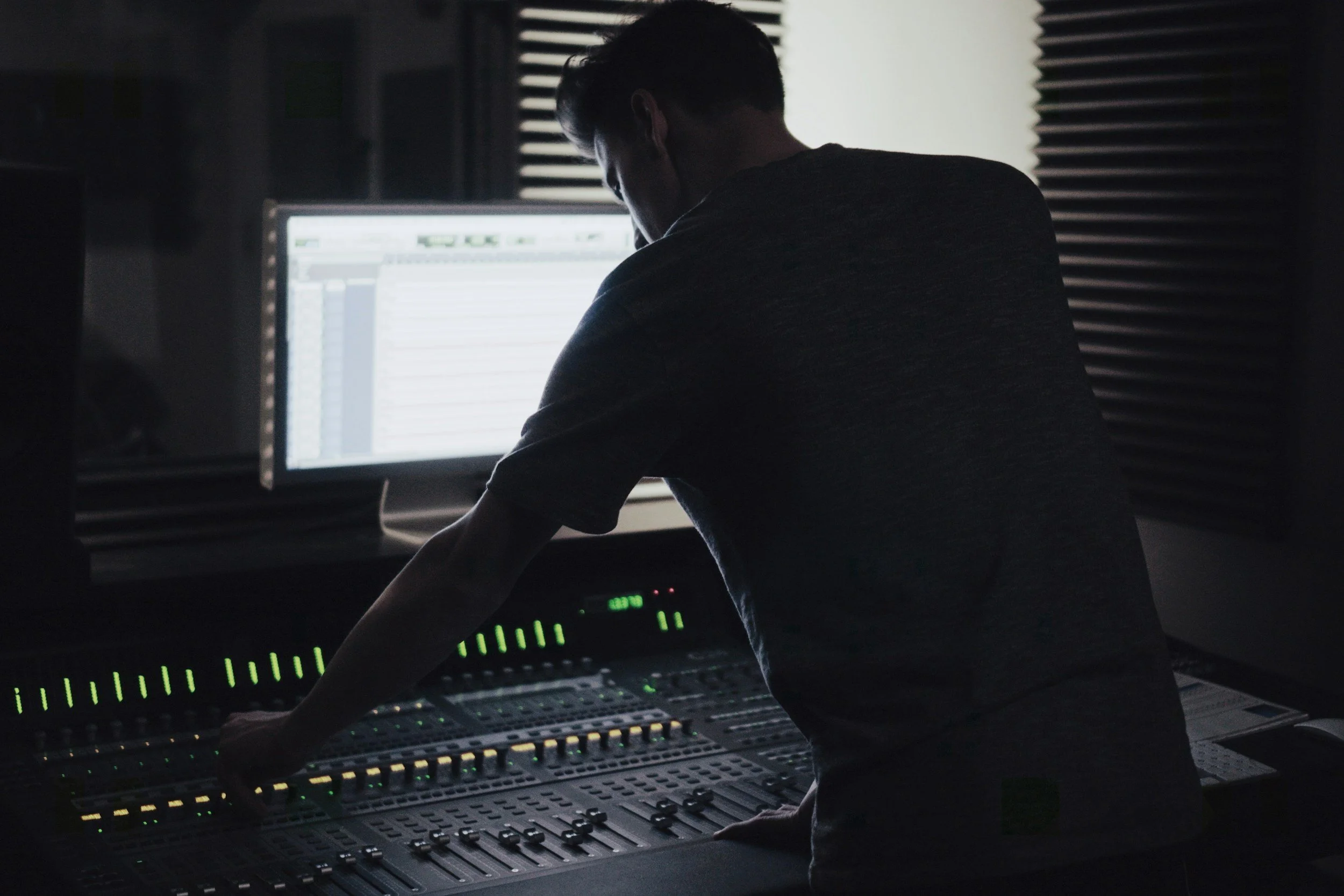 Person working on an audio mixing console in a dark room with a computer screen in the background.