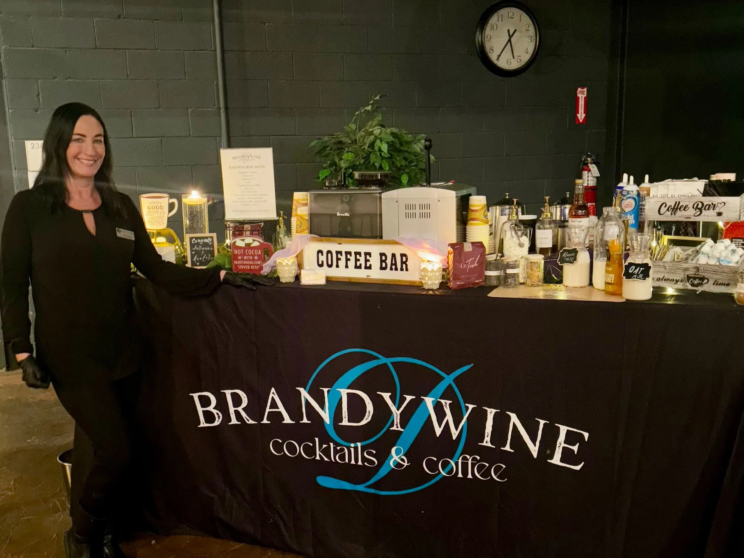 A woman wearing a black outfit and black gloves standing next to a black table with a sign that reads 'COFFEE BAR' in front of a black brick wall. The table has coffee supplies, cups, and decorative candles. There is a clock on the wall showing 6:36 
