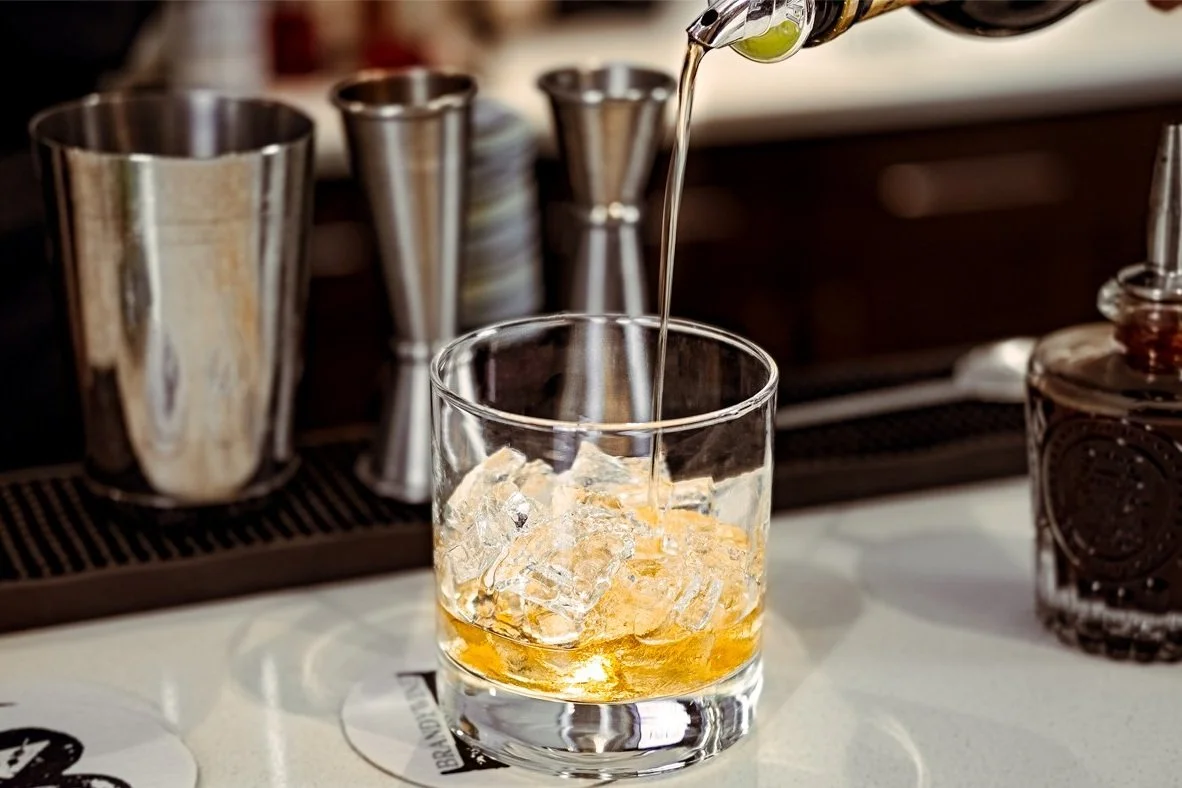 A glass with ice cubes and amber-colored liquor, being poured from a bottle, on a bar counter.