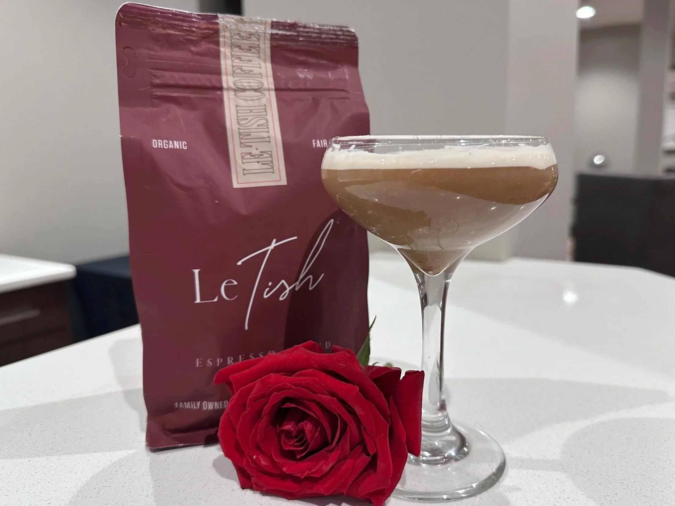 A glass of coffee or espresso with a foamy top, a maroon bag of Le Mist coffee, and a red rose on a white surface in a cozy indoor setting.