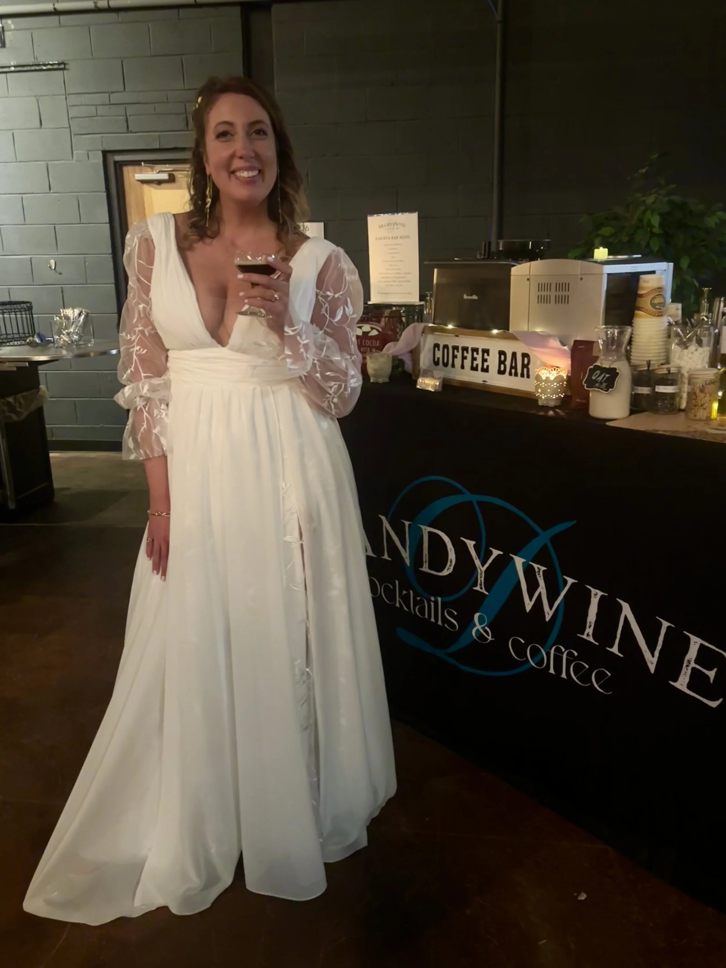 A woman in a white dress holding a glass of red wine, standing in front of a coffee bar at an event.