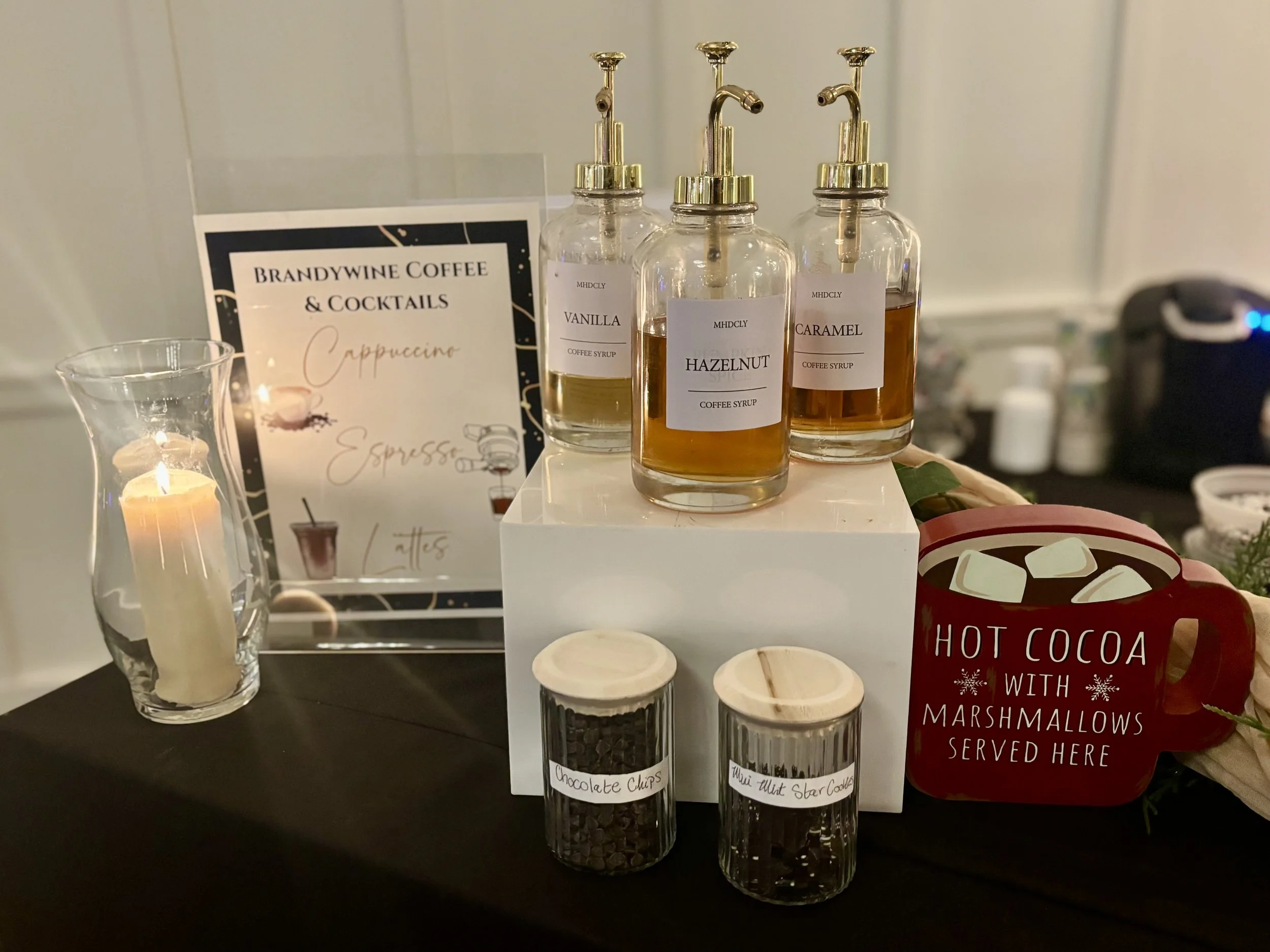 Display of three glass bottles with gold-colored pump dispensers filled with coffee syrups labeled vanilla, hazelnut, and caramel. A sign reads 'Brandywine Coffee & Cocktails' with 'Cappuccino' handwritten. Also includes a glass filled with what appe