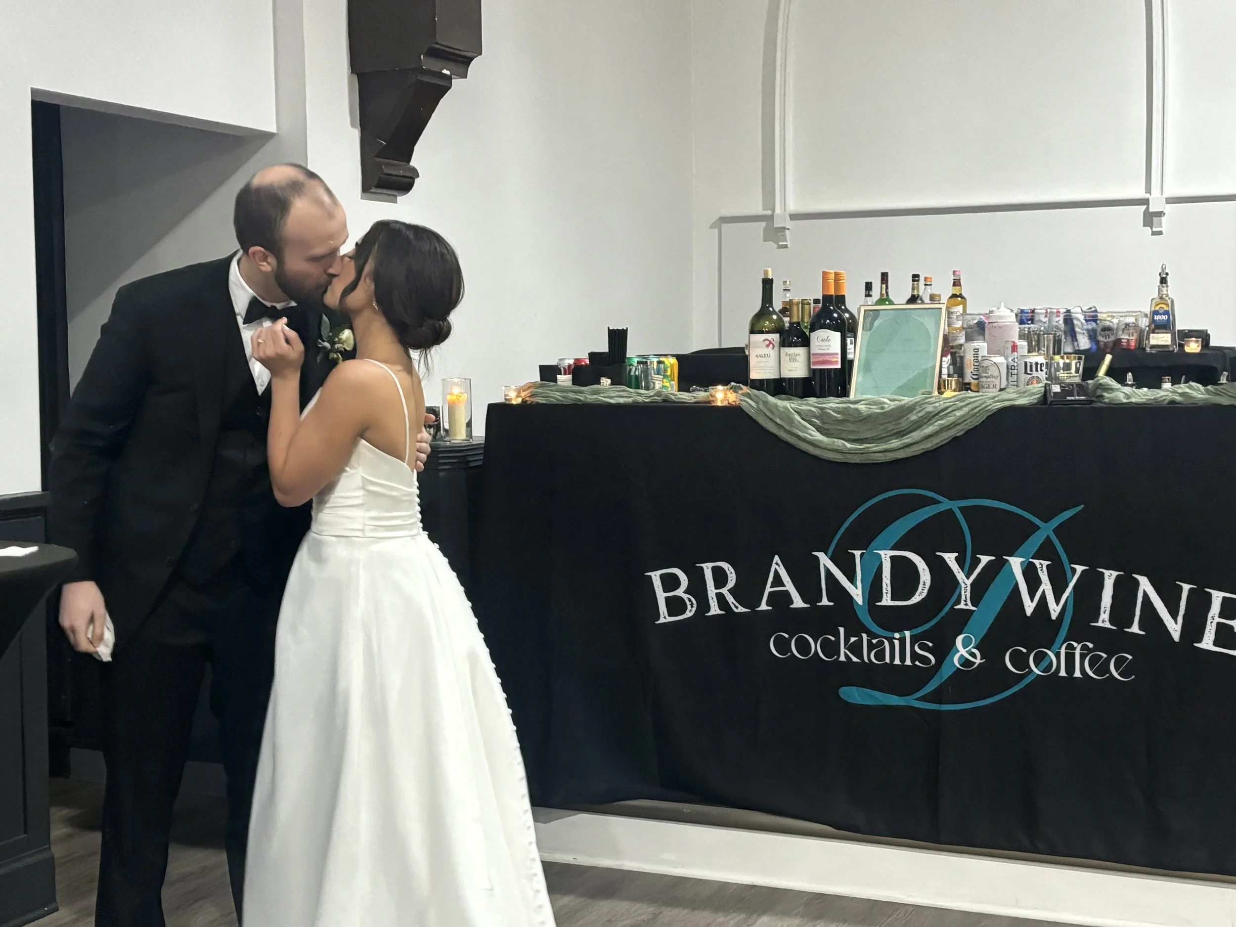 A couple dressed in formal wedding attire sharing a kiss at their wedding reception with a bar in the background.