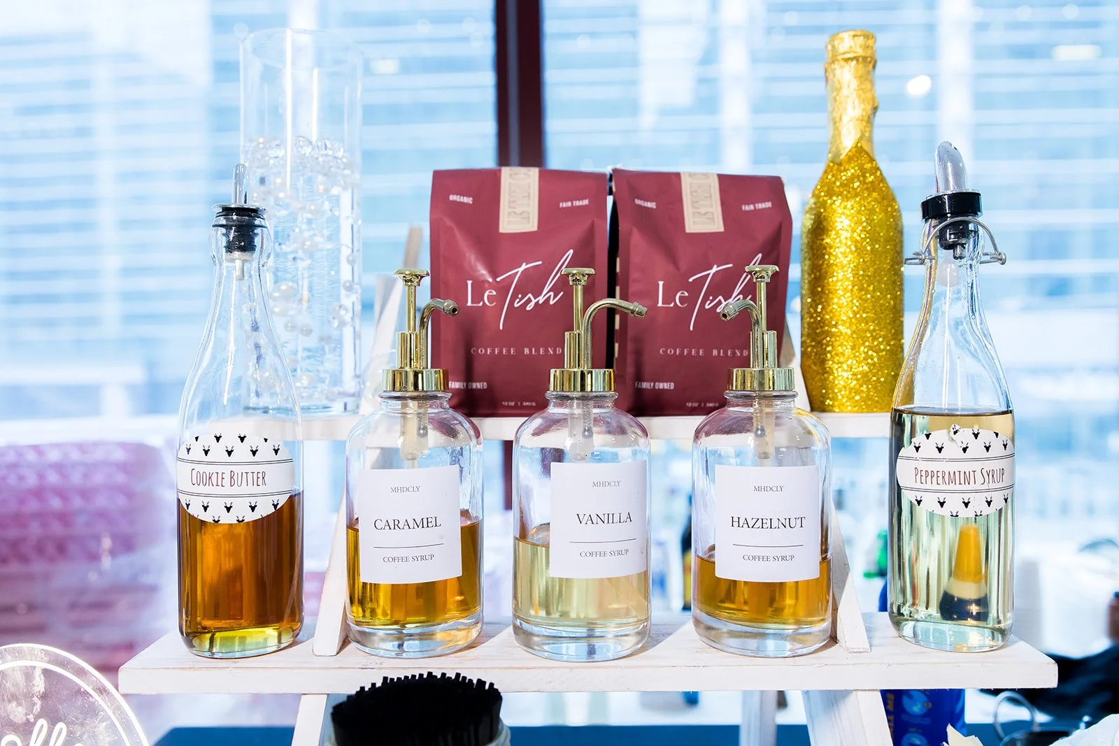 Display of flavored coffee syrups in glass bottles with pour spouts, labeled caramel, vanilla, hazelnut, and peppermint syrup, set on a white stand with coffee bean bags labeled 'Le Tish' in the background.