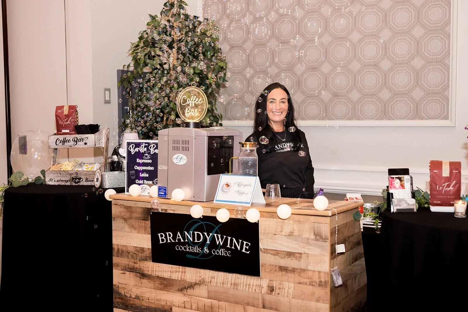 A woman standing behind a wooden coffee and cocktail bar at an event, smiling. The bar has a sign that reads 'BRANDYWINE cocktails & coffee.' There are various coffee packages, a bubble machine, and a sign with coffee drinks listed. Decorative string lights are draped across the front of the bar, and a Christmas tree with bubbles is visible in the background.