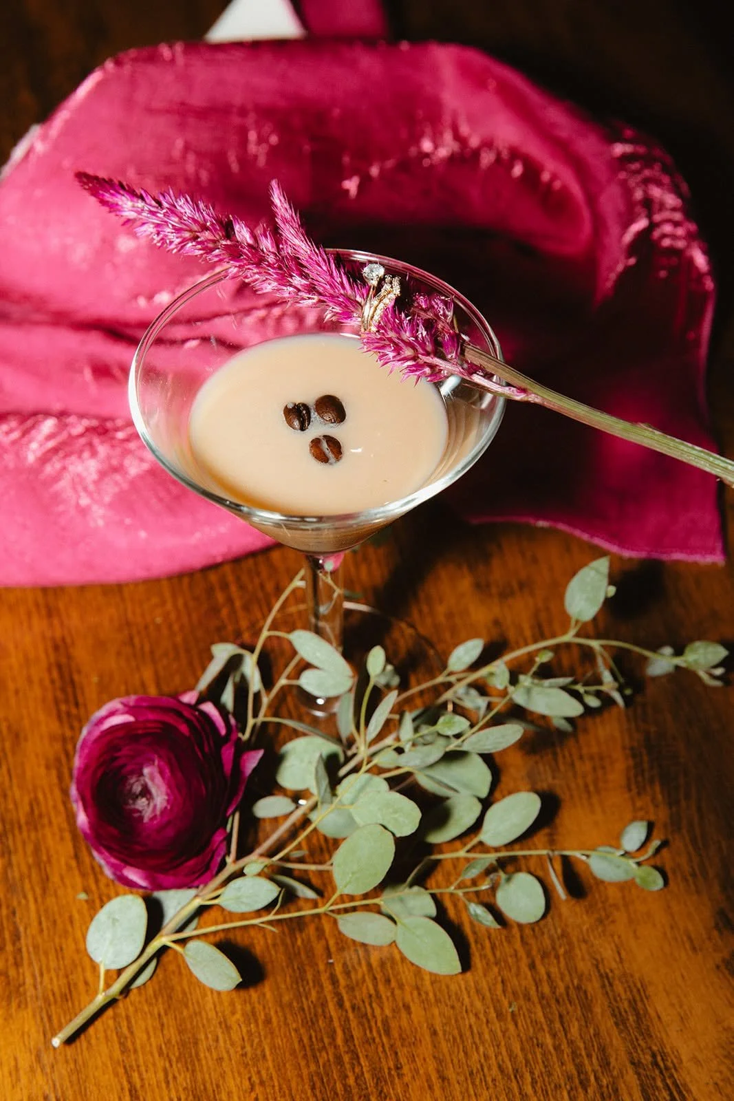 A glass of iced coffee garnished with coffee beans and a pink flower, set on a wooden table with a pink cloth and green leaves surrounding it.
