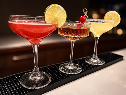 Three cocktails on a bar counter, each garnished with lemon slices and cherries, in different colored glasses.