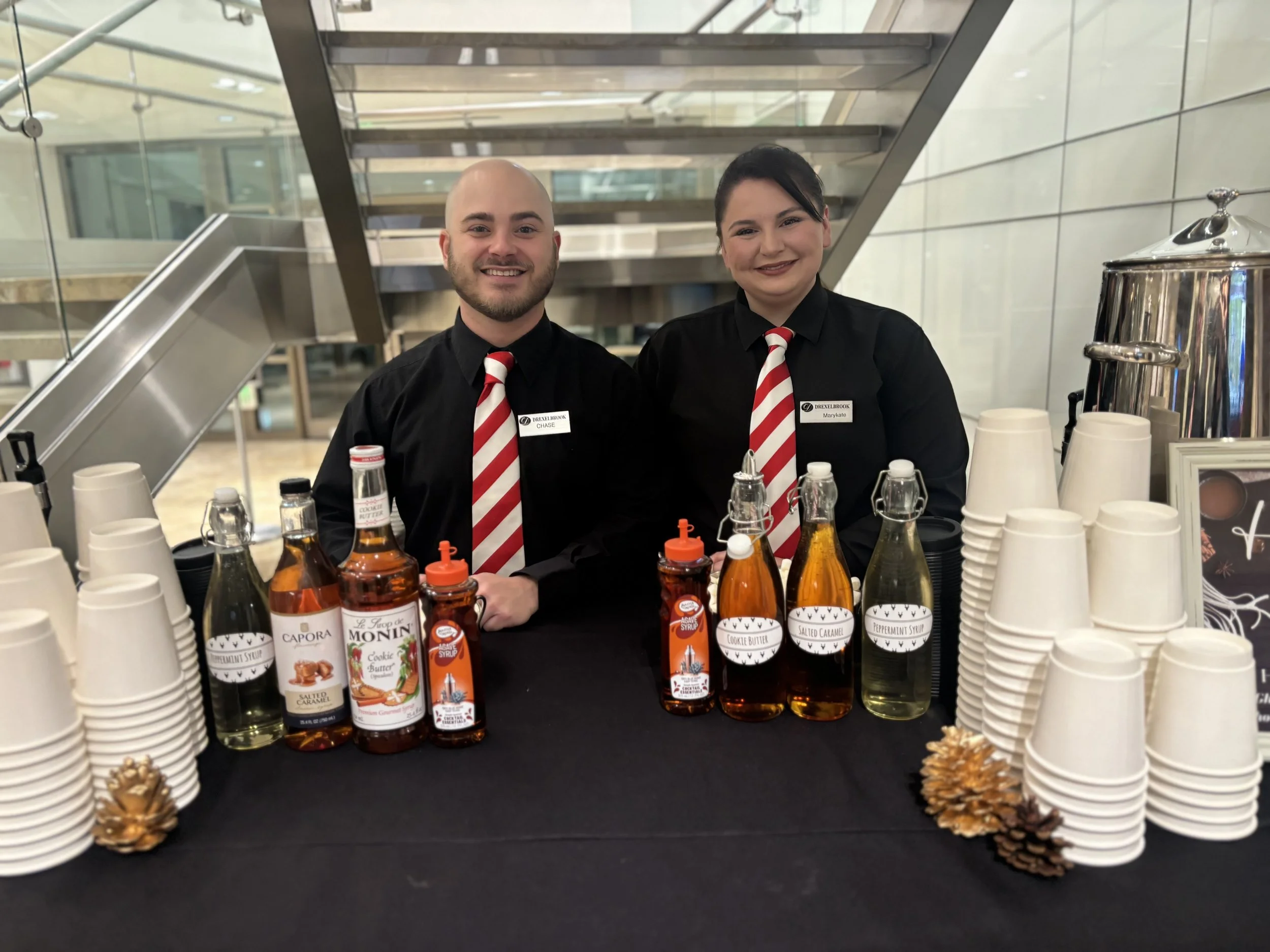 Two smiling employees, a man with a shaved head and a woman with dark hair, wearing black shirts and red-and-white striped ties, stand behind a table with drink ingredients and cups at a coffee shop or cafe. The background features a modern interior 