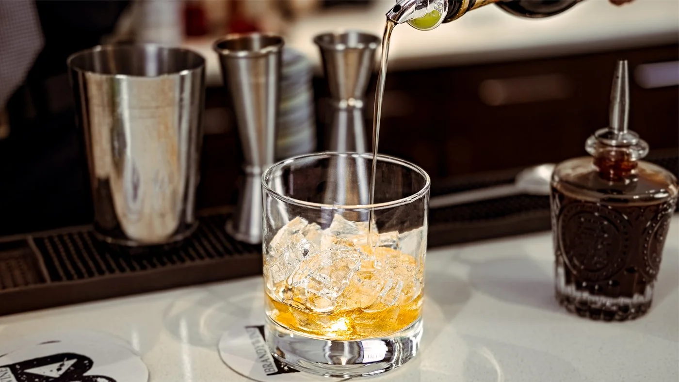 A glass with ice cubes being filled with amber-colored liquid from a bottle, situated on a bar counter with cocktail shakers and a syrup dispenser in the background.