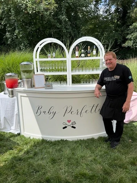 Outside beverage bar with a white counter and arch decoration, labeled 'Baby Perfect,' with a smiling man in a black shirt and apron standing next to it.