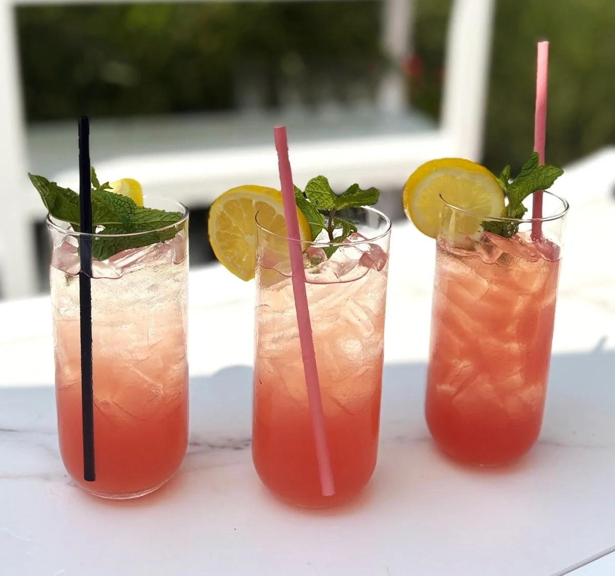 Three tall glasses of pink lemonade garnished with lemon slices and fresh mint leaves, each with a straw, placed on a white surface outdoors.