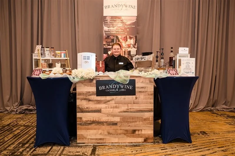 A woman standing behind a booth labeled 'Brandywine Cocktails & Coffee,' with a variety of drinks and items displayed on the table. The booth is set up indoors with beige curtains in the background and a patterned carpet on the floor.