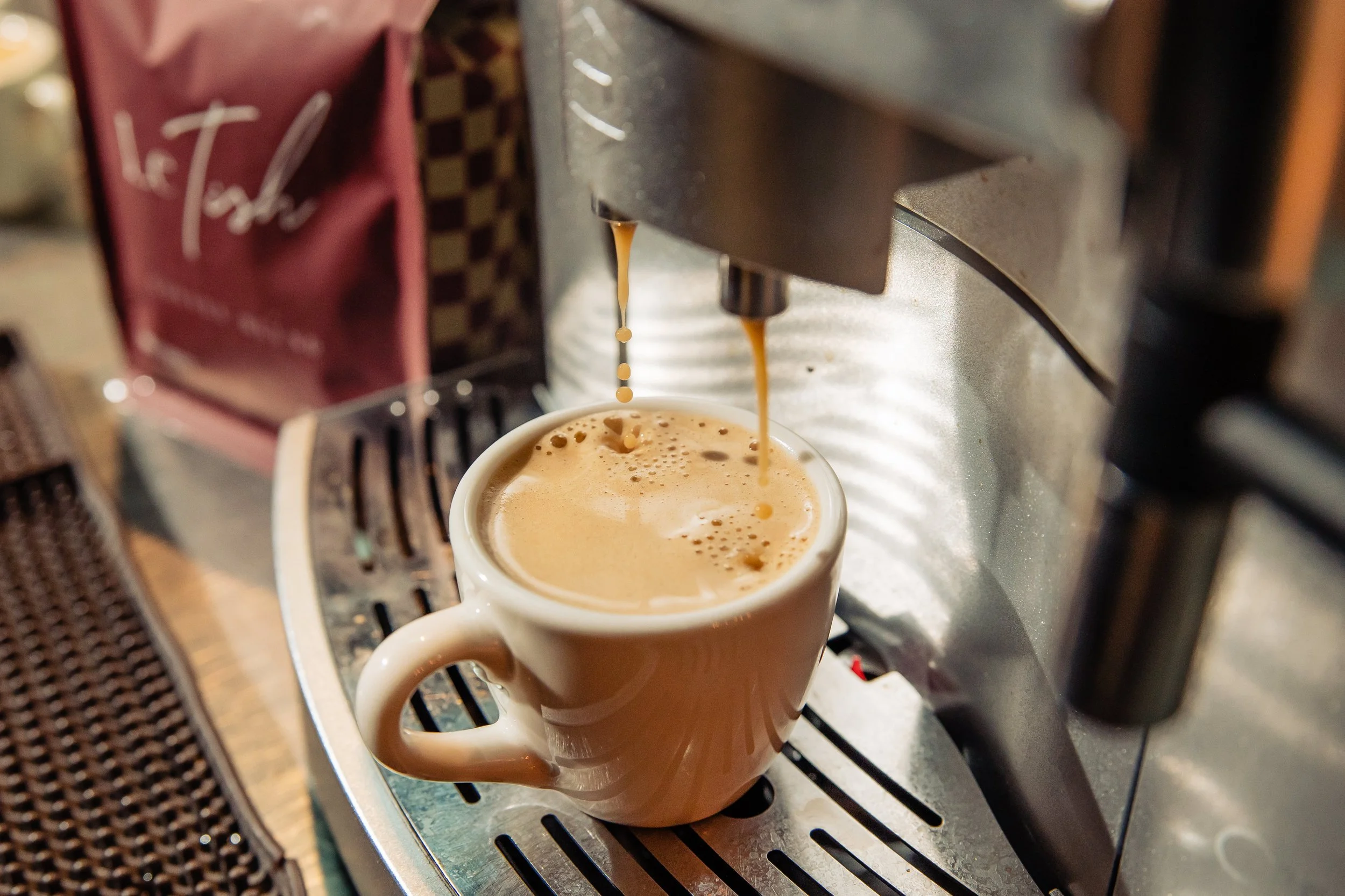 Close-up of a coffee machine brewing coffee into a ceramic mug. The mug is white with a handle, and the coffee is light brown with foam. The coffee machine has metallic and black components, with coffee dripping from the spout into the mug.