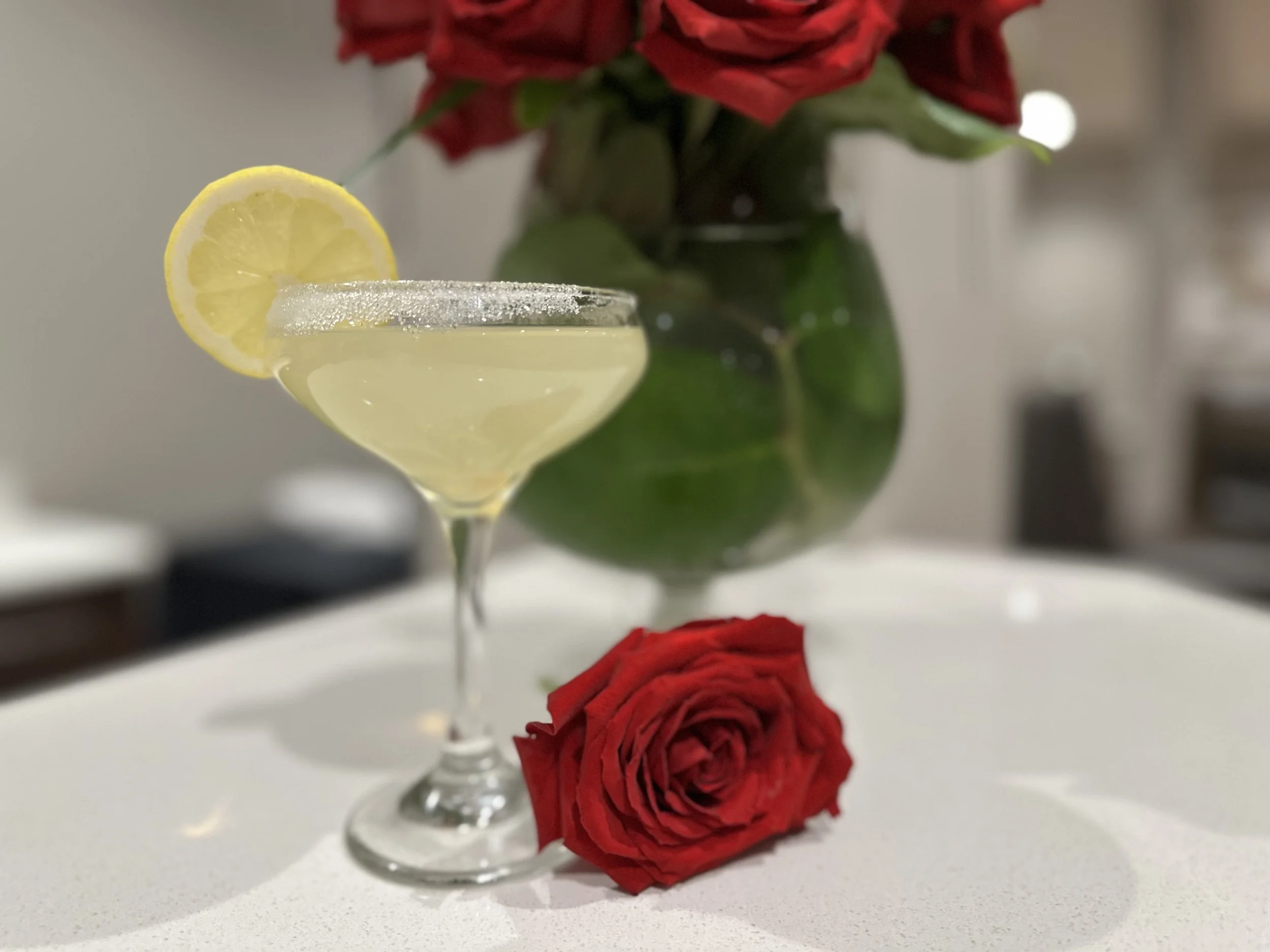 A glass of lemonade garnished with a lemon slice sits on a white surface. Behind it, a bouquet of red roses in a green vase is visible, with some roses also lying on the surface.