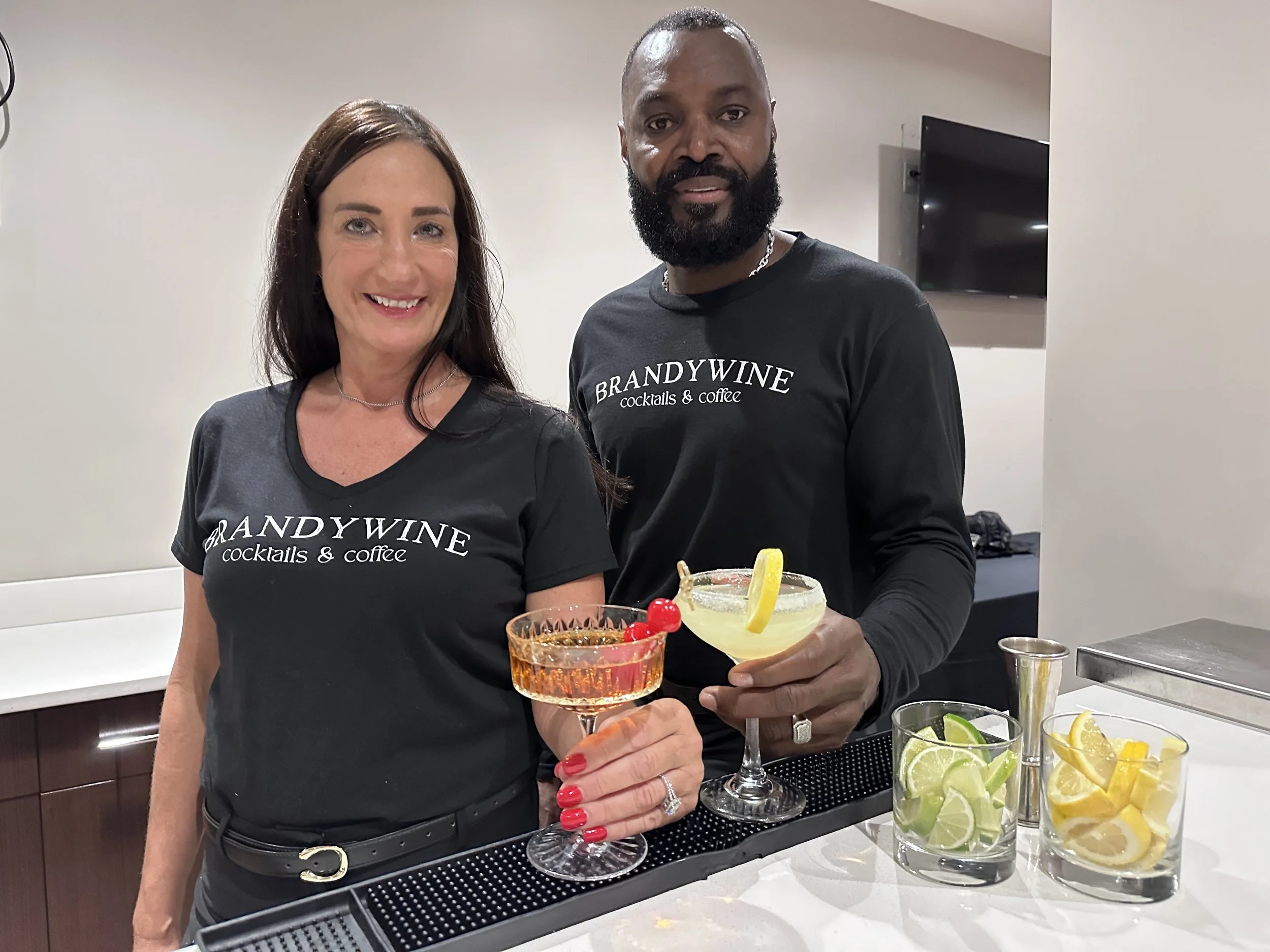 Two people standing behind a bar counter, holding cocktails with lemon and cherry garnishes. They are both smiling and wearing matching branded T-shirts that say "BRANDYWINE cocktails & coffee." The bar area has various garnishes including lemons and