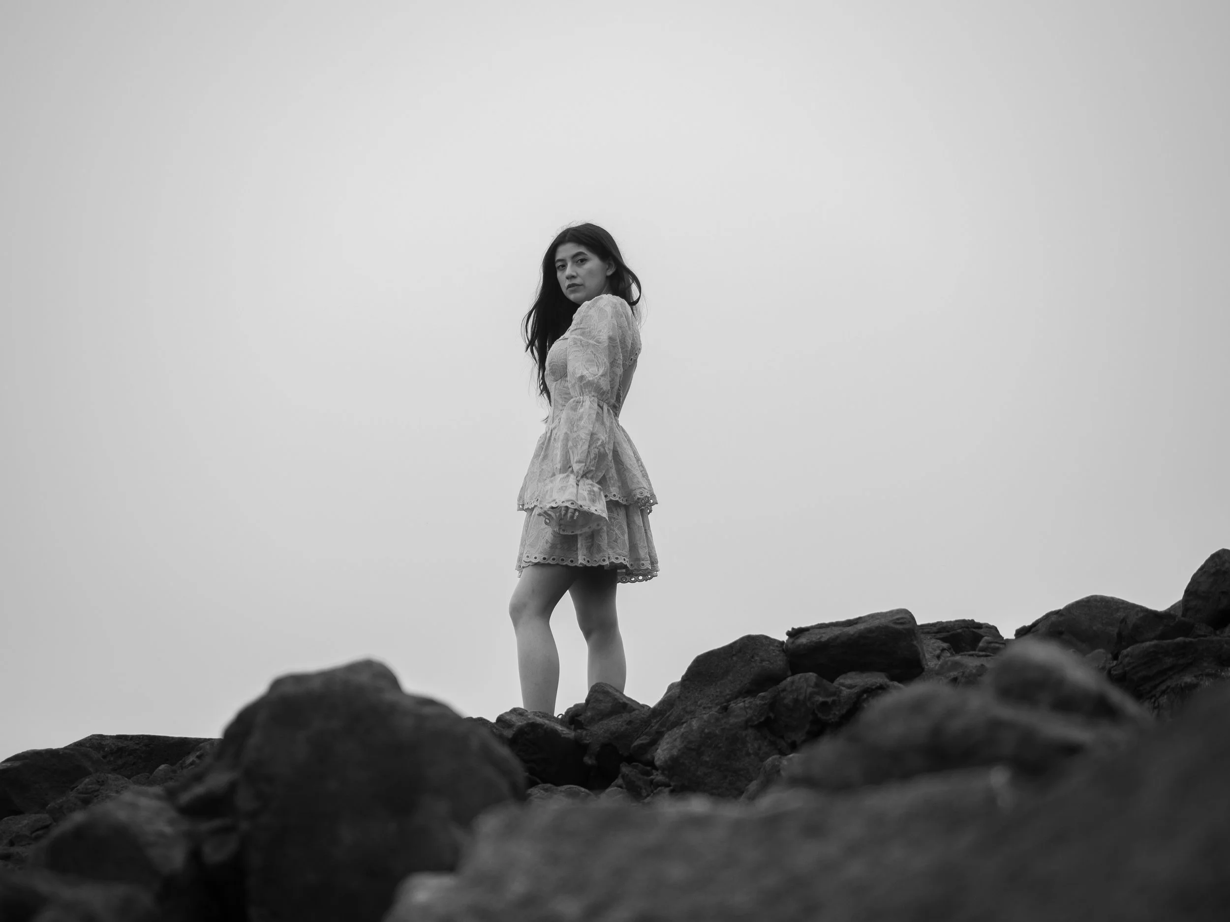 Schwarz-Weiß-Foto einer Frau im Kleid, die auf Felsen steht, mit bewölktem Himmel im Hintergrund.