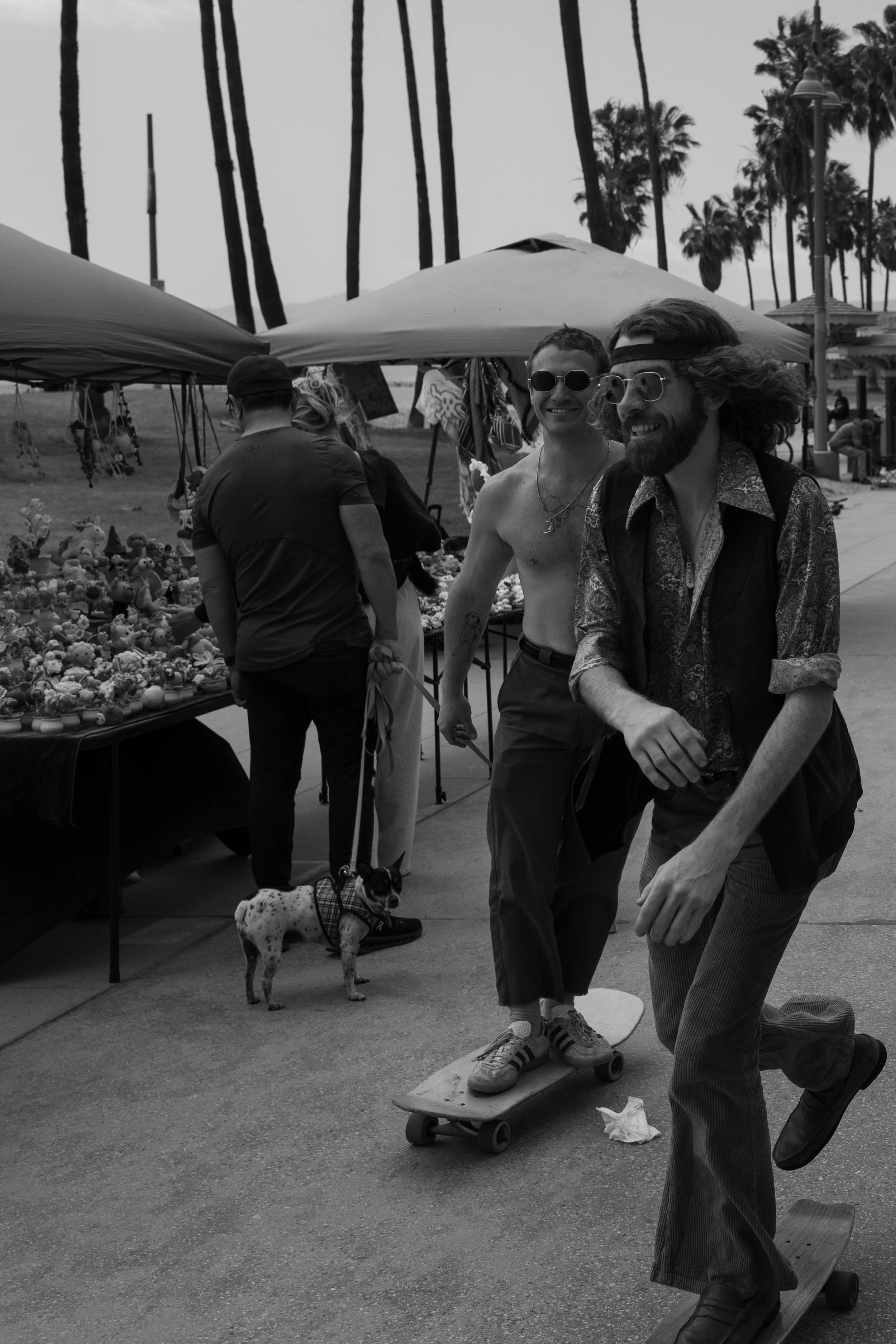 Zwei Männer, einer nackt mit Sonnenbrille, fährt auf einem Skateboard, während der andere mit langen Haaren und Bart ebenfalls auf einem Skateboard fährt. Im Hintergrund ein Marktstand mit Puppen und anderen Gegenständen, Palmen und mehrere Menschen.