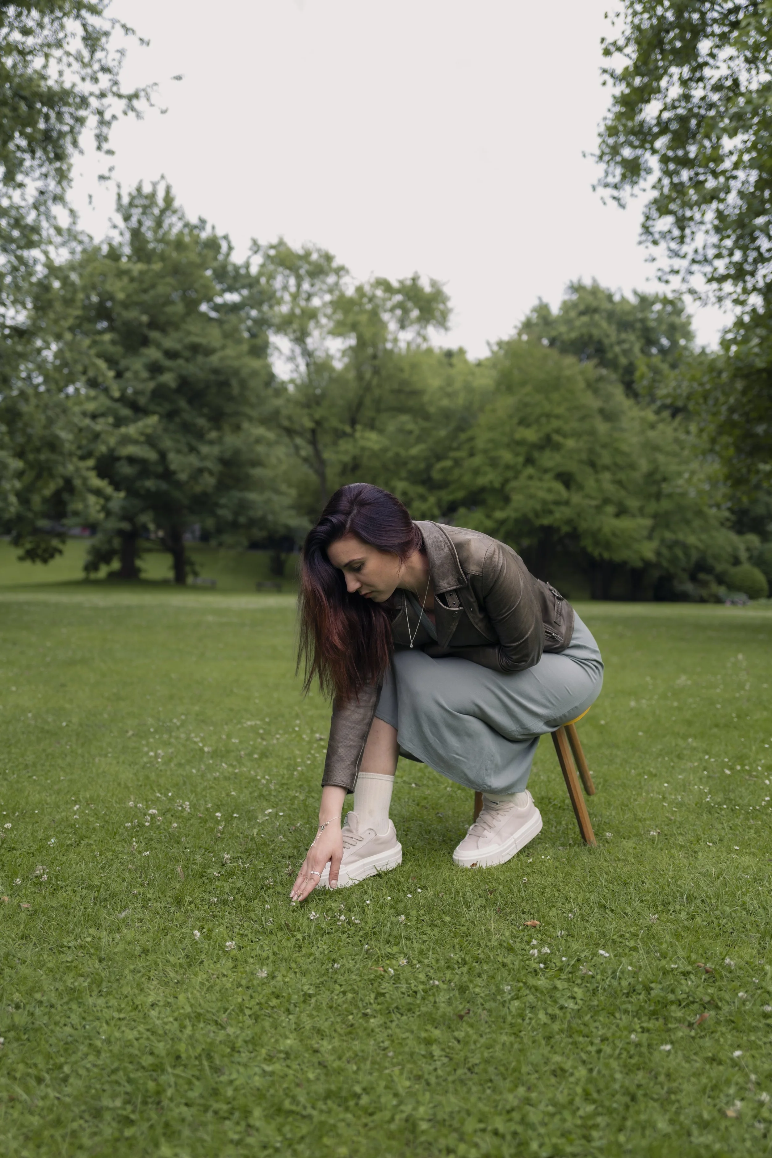 Junge Frau sitzt auf einem Stuhl im Park, beugt sich nach vorne und berührt den Boden mit ihrer Hand, während sie auf grünem Rasen steht.