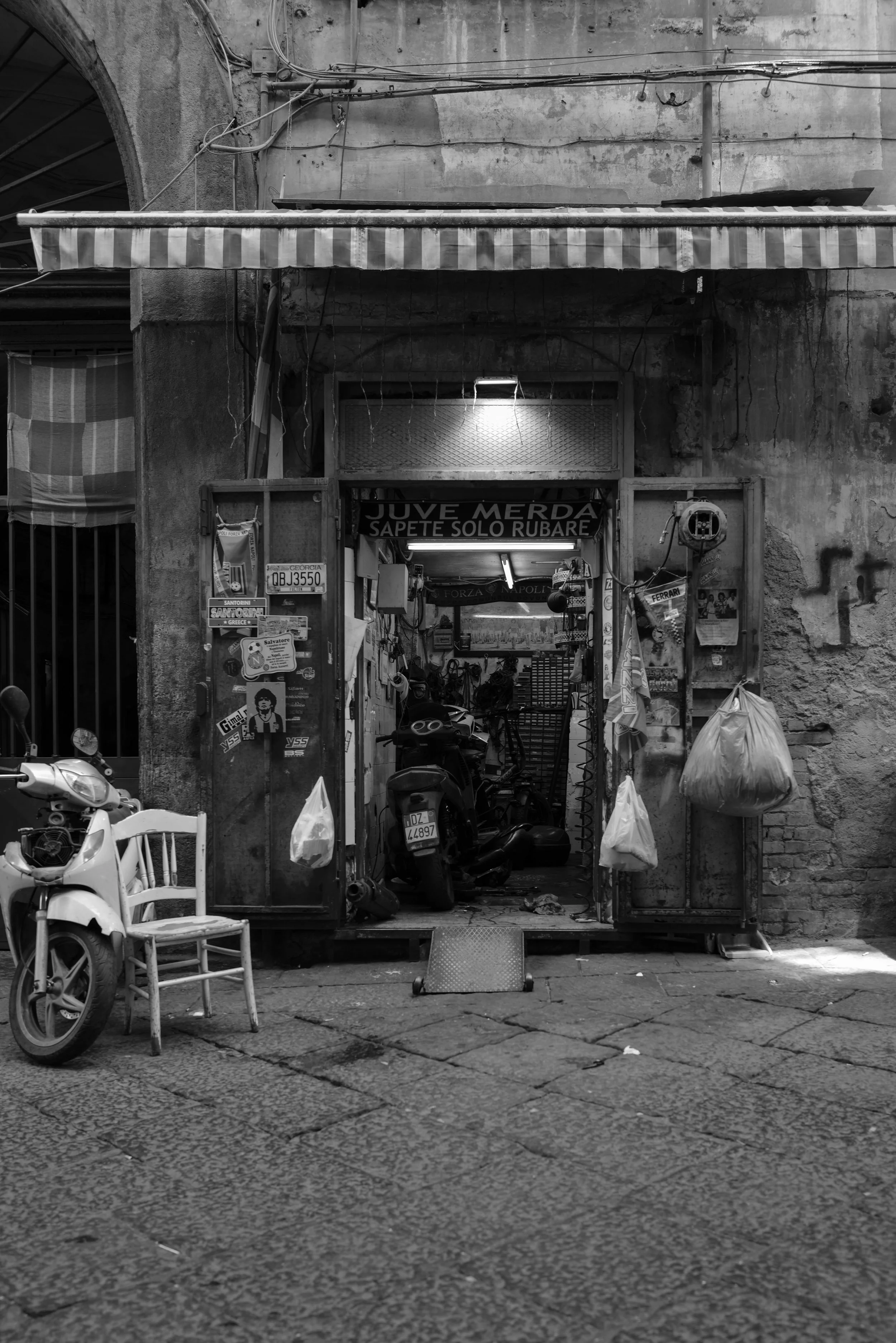 Schwarz-Weiß-Foto eines kleinen, rustikalen Ladens in einer Stadt, mit einem Motorrad und einem Elektro-Scooter vor dem Eingang. Die Ladenfront ist alt mit Straßenschildern und Aufklebern verziert. Ein weißer Stuhl steht neben dem Eingang, und Plasti