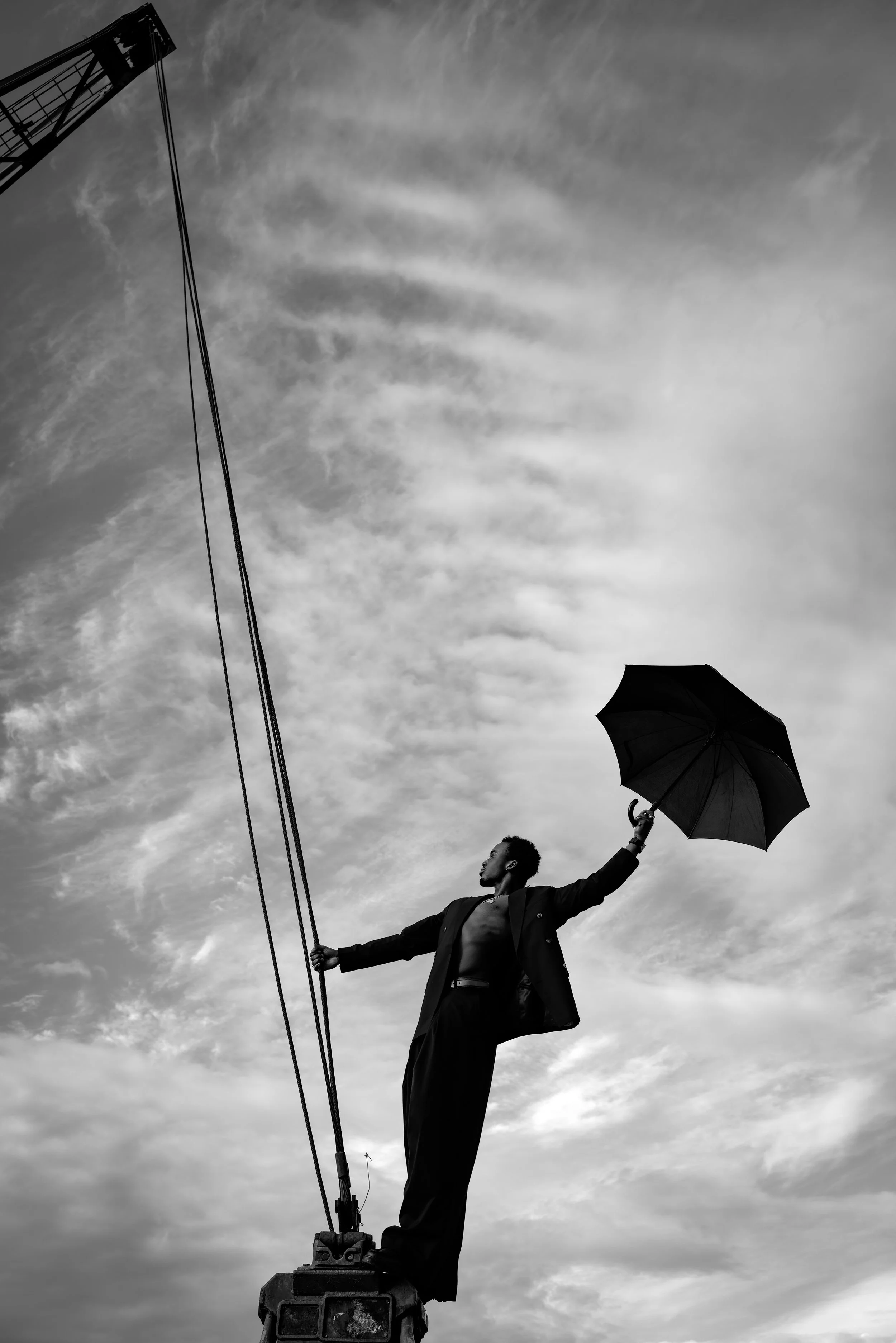 Schwarze Silhouette eines Mannes auf einer Begrenzung, hält einen schwarzen Regenschirm in der Hand, während er auf einer erhöhten Plattform steht, gegen einen Himmel mit Wolken im Hintergrund, schwarz-weiße Fotografie.