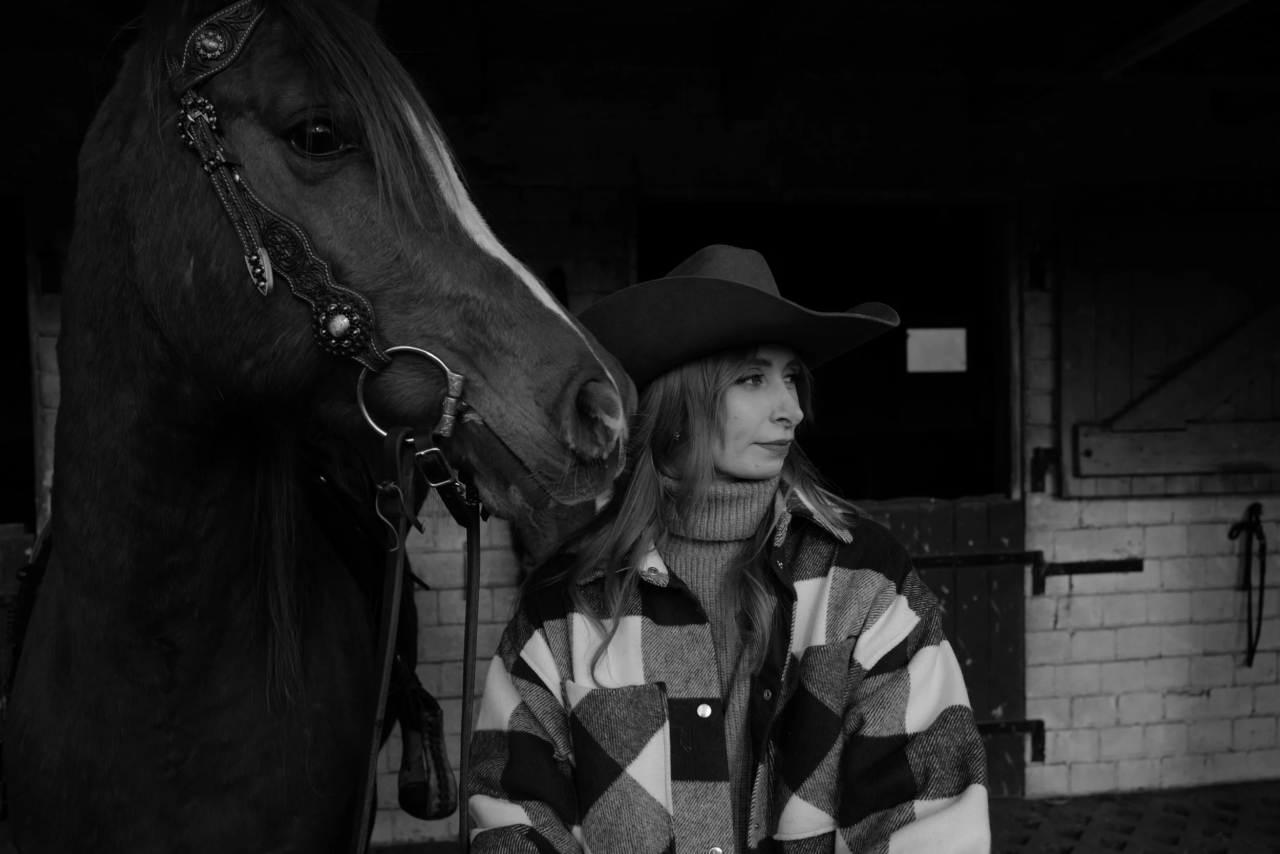 Frau mit Cowboyhut neben Pferd in Schwarz-Weiß-Optik vor einem Stall stehend.