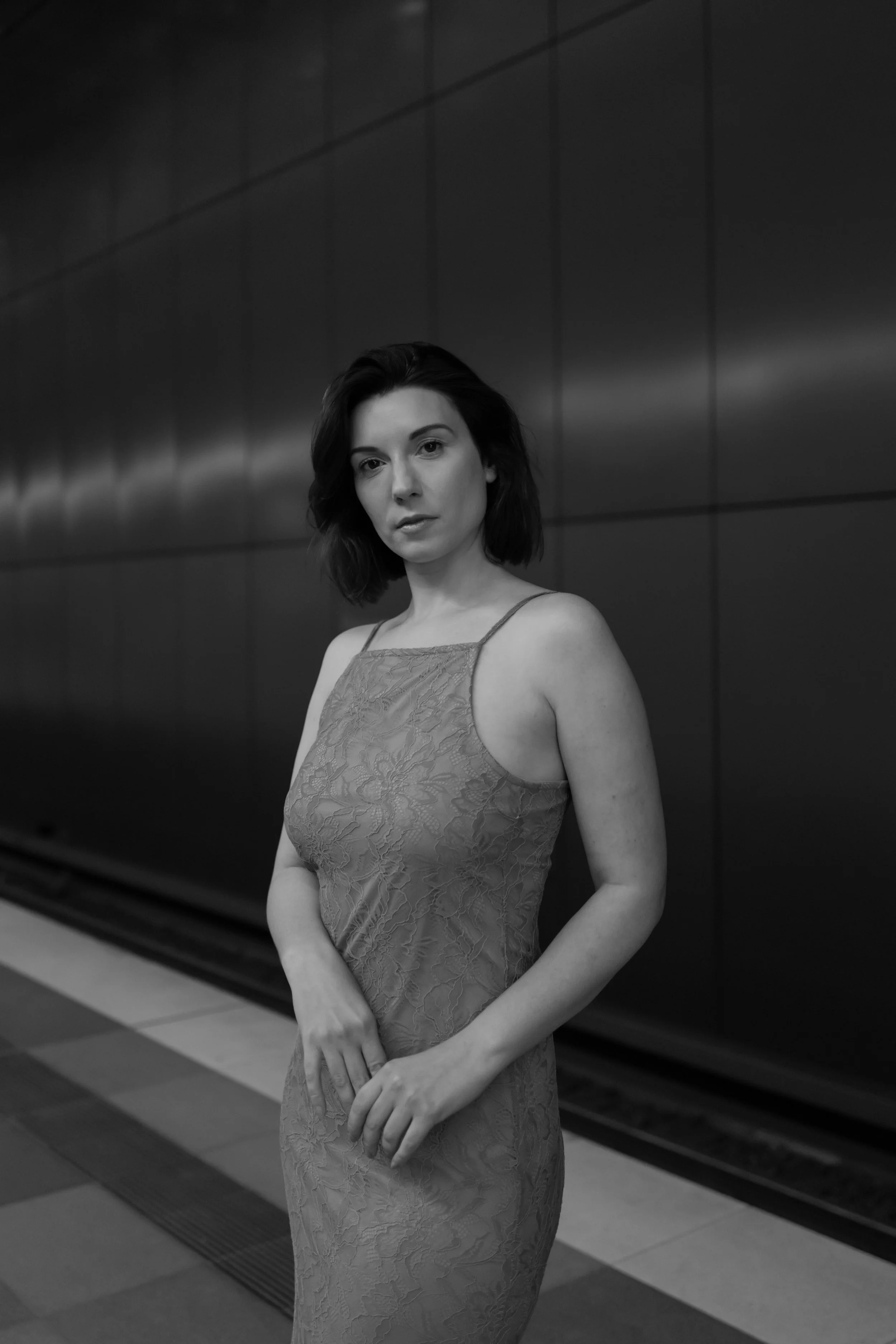 Schwarz-Weiß-Foto einer jungen Frau in einem eleganten Kleid, die vor einer dunklen Wand steht, mit einem nachdenklichen Ausdruck.