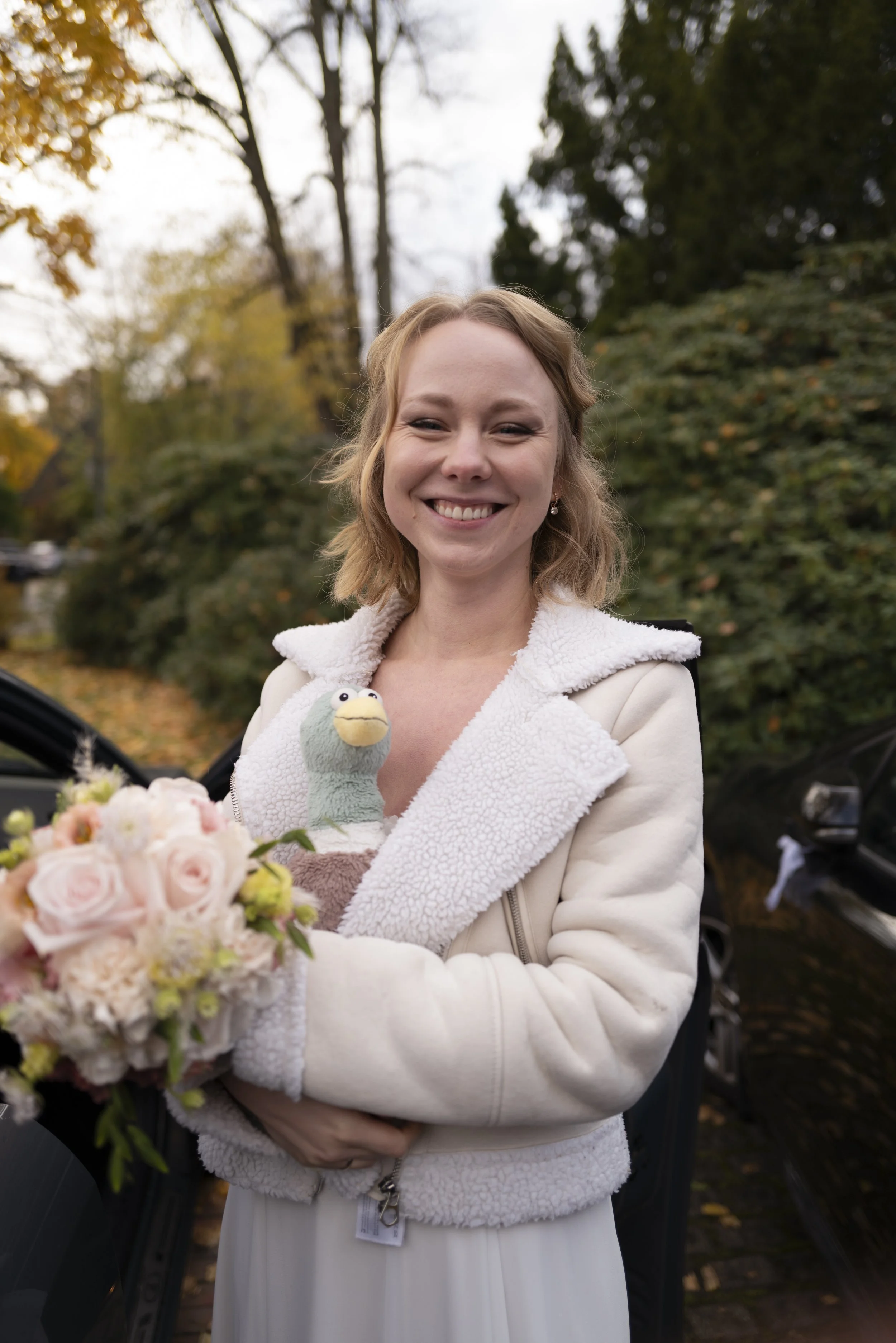Eine lachende Frau mit roten Haaren hält einen Blumenstrauß mit rosa Rosen und weißen und grünen Blättern. Sie trägt einen weißen Mantel mit Fellkragen und steht im Freien vor Bäumen mit Herbstlaub.