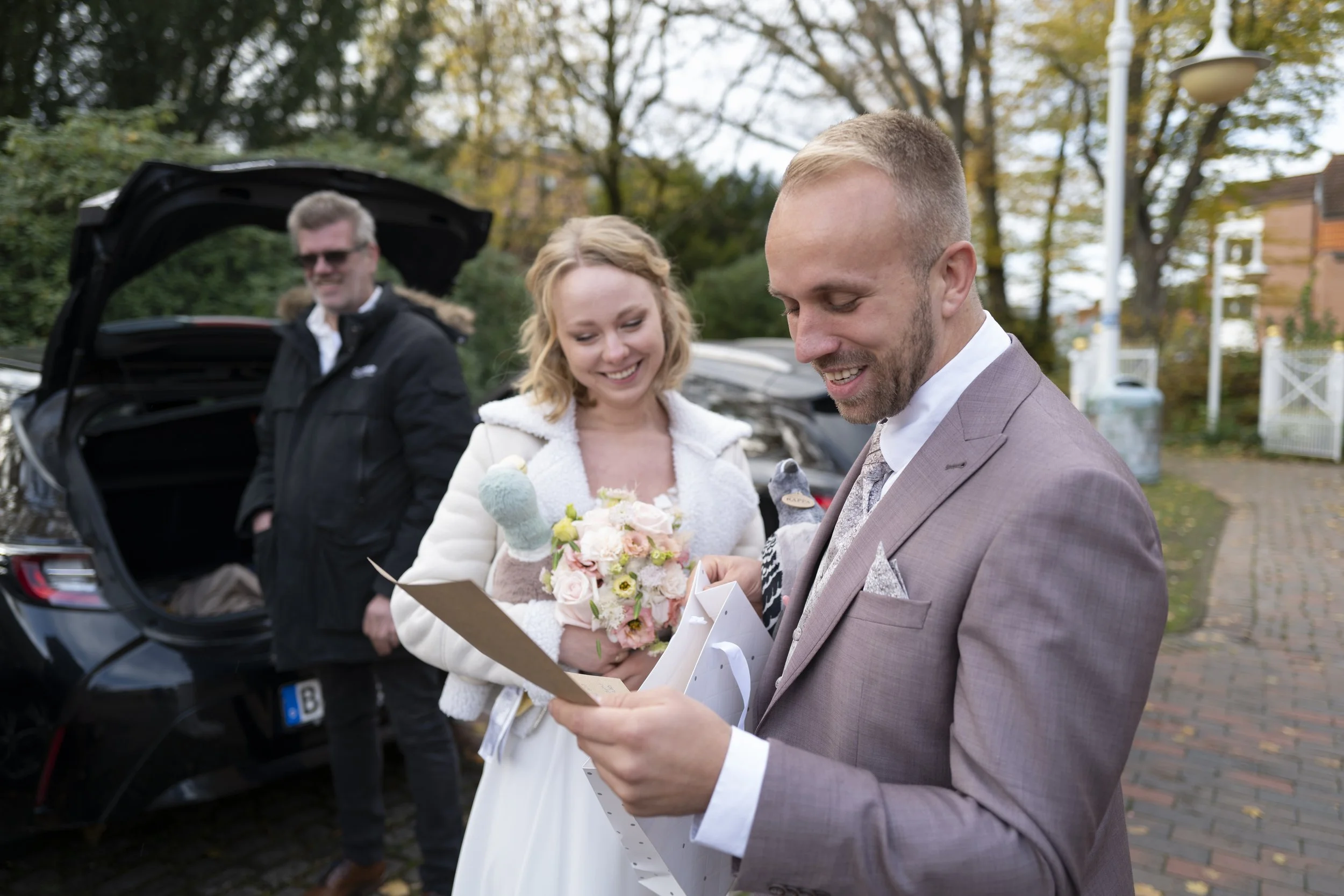Ein Mann in einem Anzug erhält ein Geschenk und einen Blumenstrauß von einer Braut bei einer Hochzeit im Freien, während eine Person im Hintergrund die Motorhaube eines Autos öffnet.