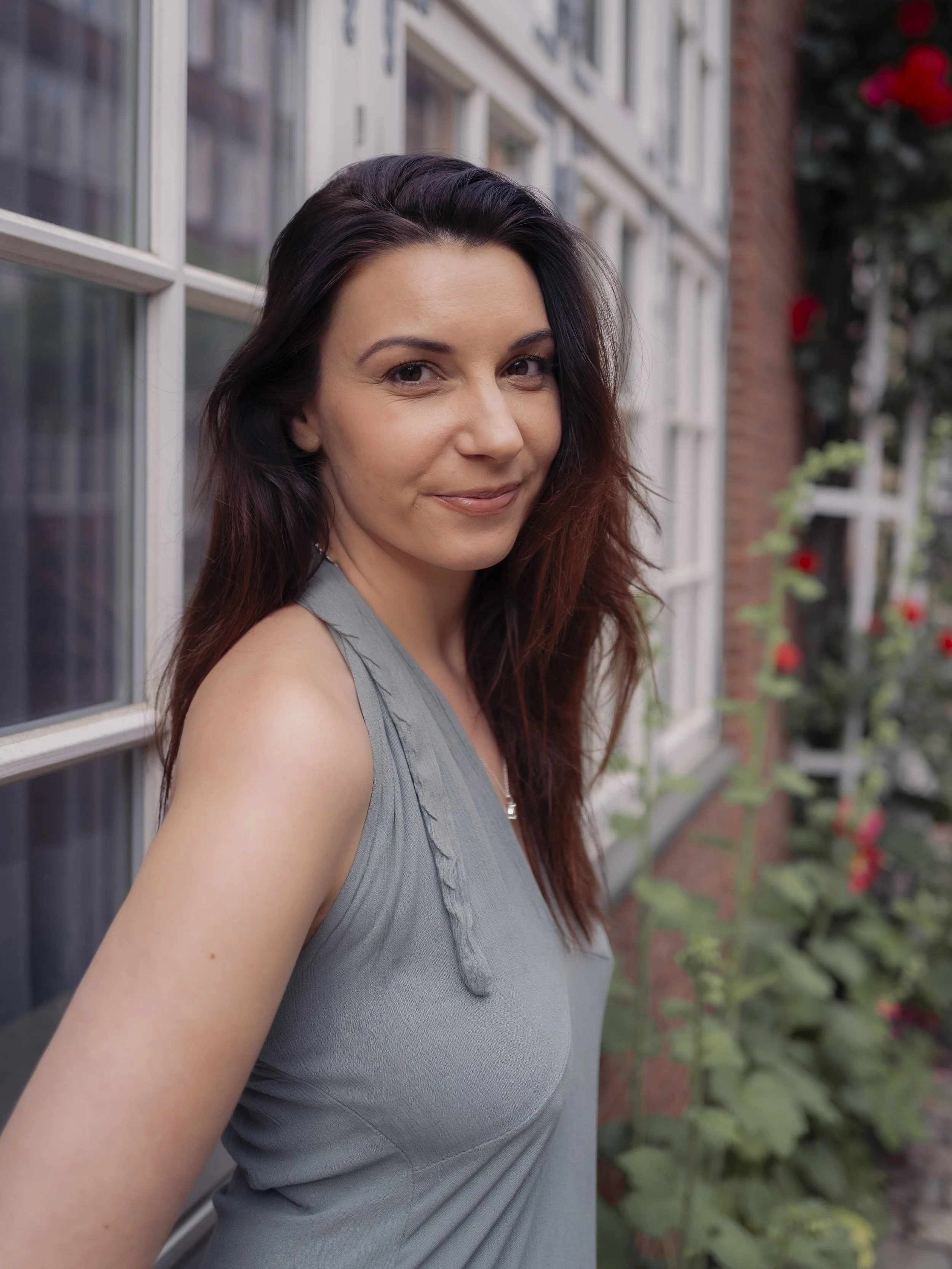 Portrait einer jungen Frau mit dunklen, gewellten Haaren vor einem Fenster und mit grünen Pflanzen im Hintergrund.