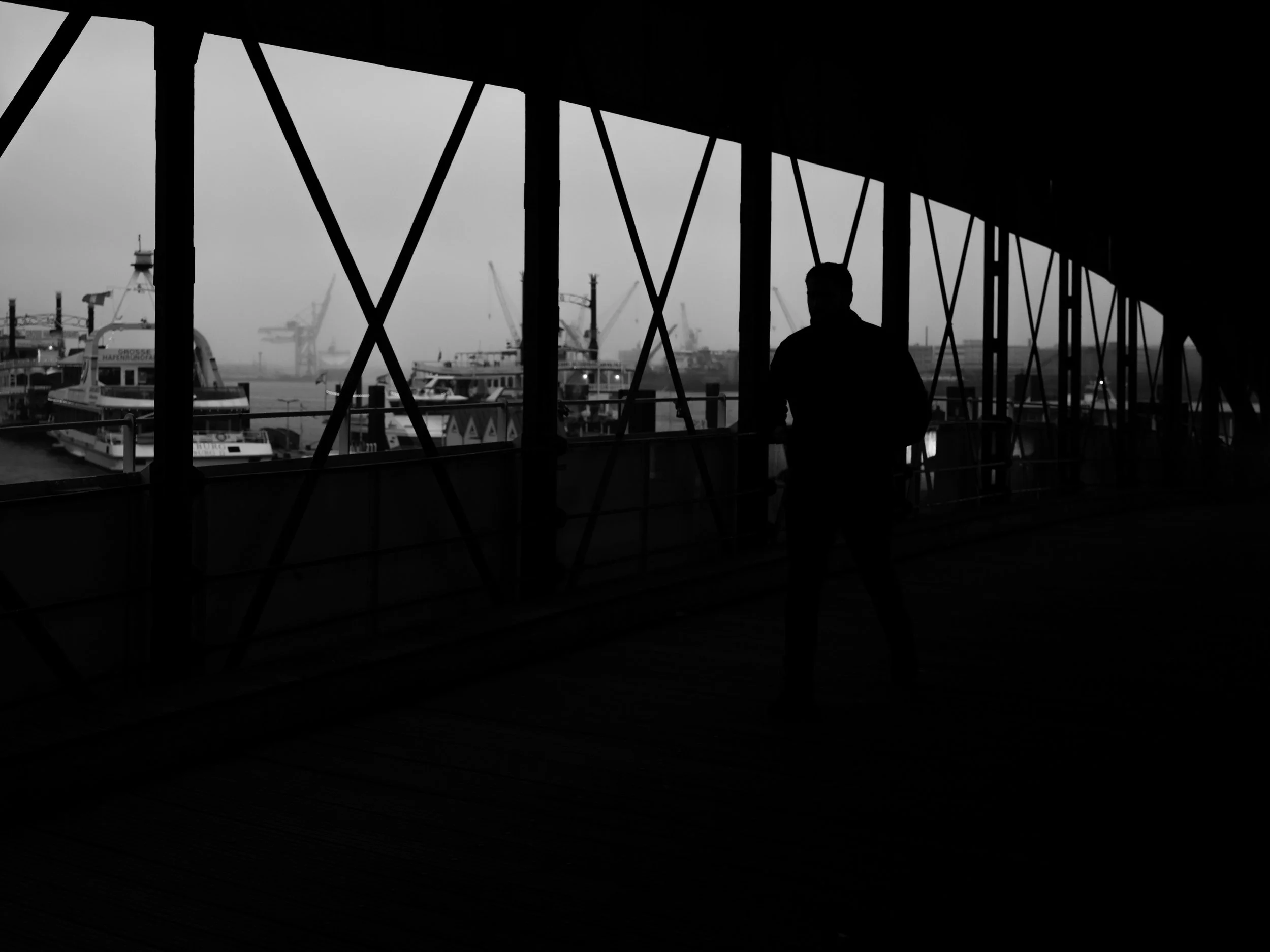 Schwarz-Weiß-Fotografie eines Hafenblicks durch Fenster mit Gitterstreben. Silhouette einer Person im Vordergrund. Im Hintergrund sind Schiffe und Kräne sichtbar.