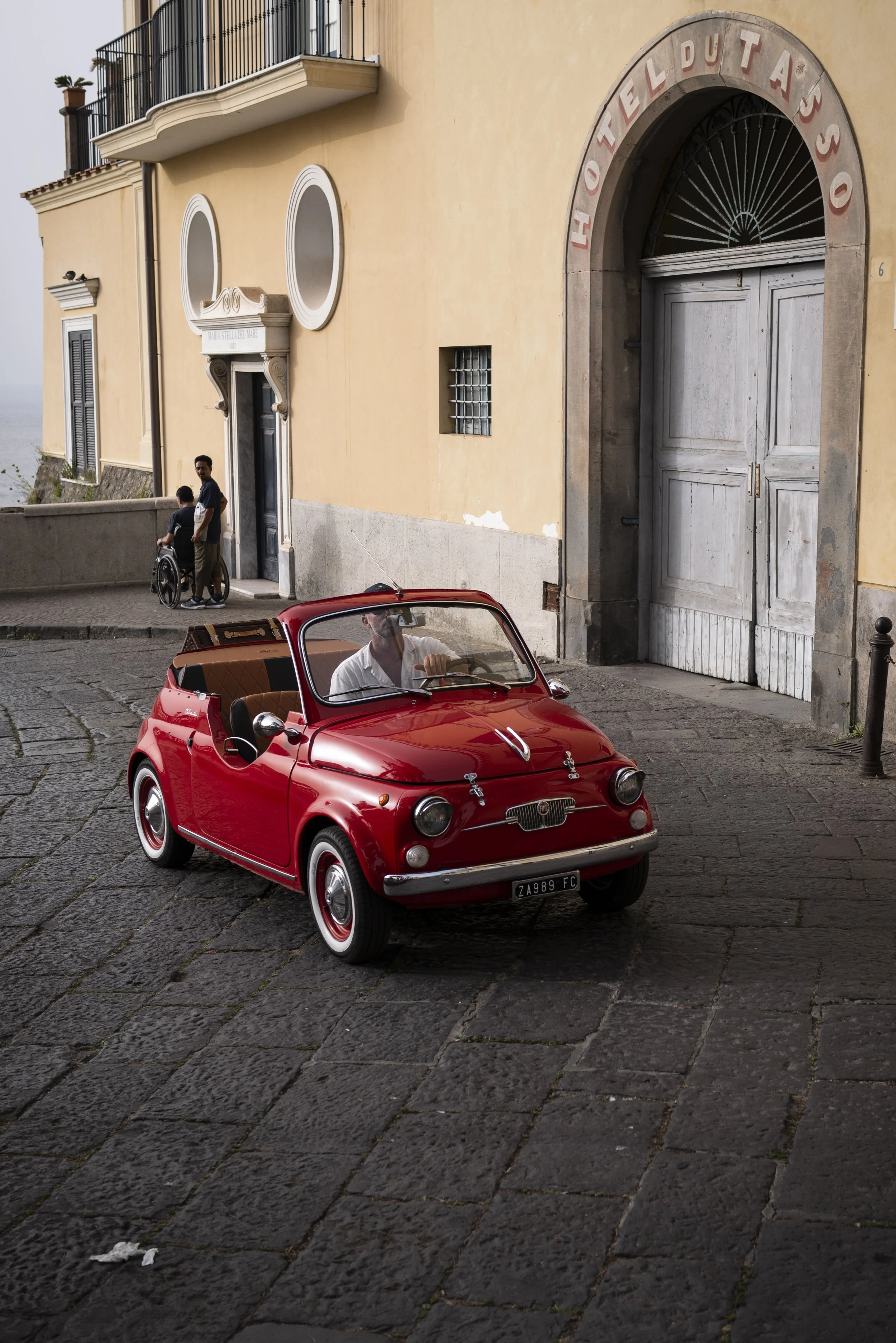 Rotes Cabrio Fiat 500 auf einer gepflasterten Straße vor einem Gebäude mit der Aufschrift 'HOTEL DU TASSO' und zwei Personen in der Nähe.