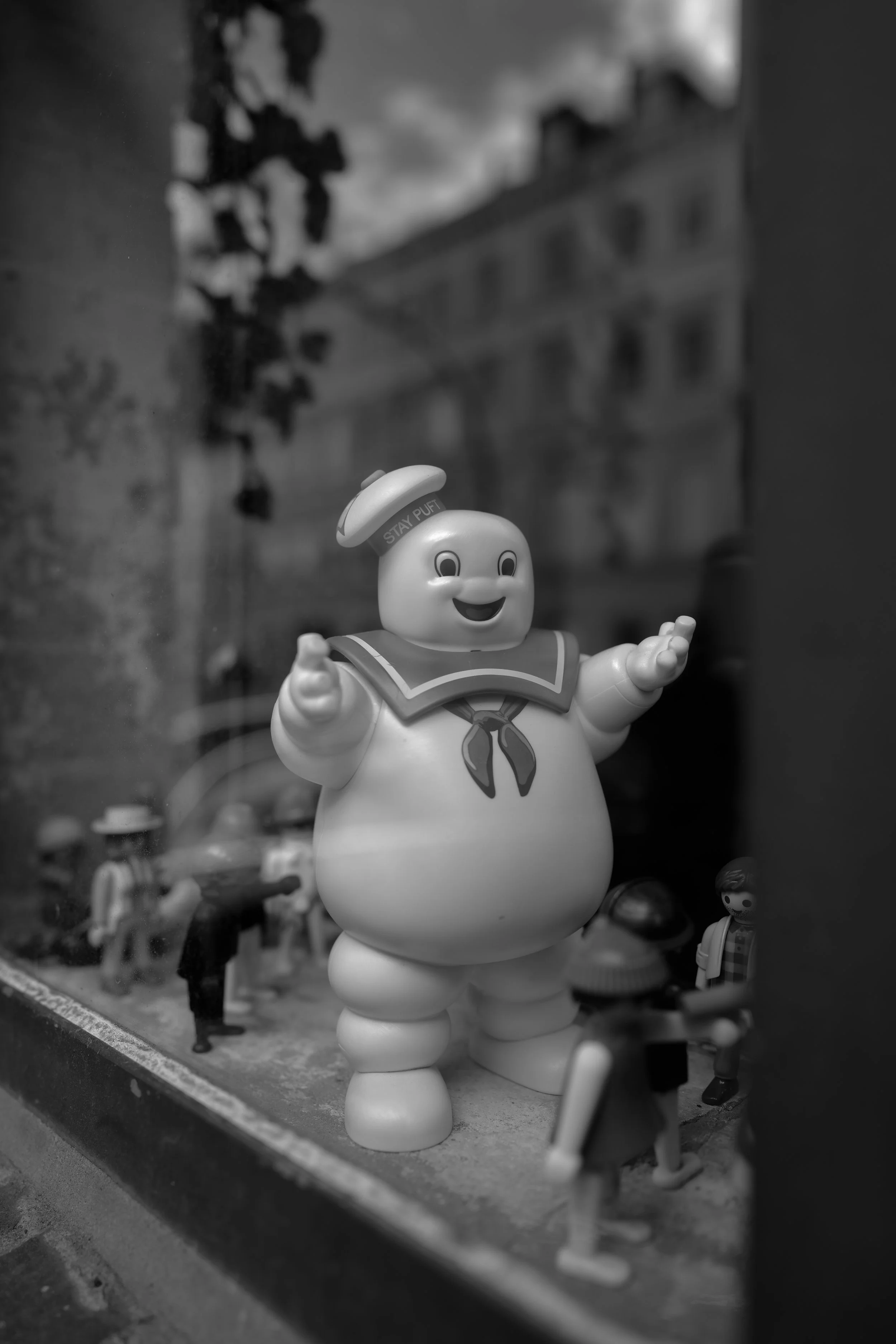 Ein figurineähnliches Spielzeug eines freundlichen Geist mit einem Hut, das auf einem Fensterbrett steht, umgeben von weiteren kleinen Figuren, mit Gebäuden und Wolken im Hintergrund.