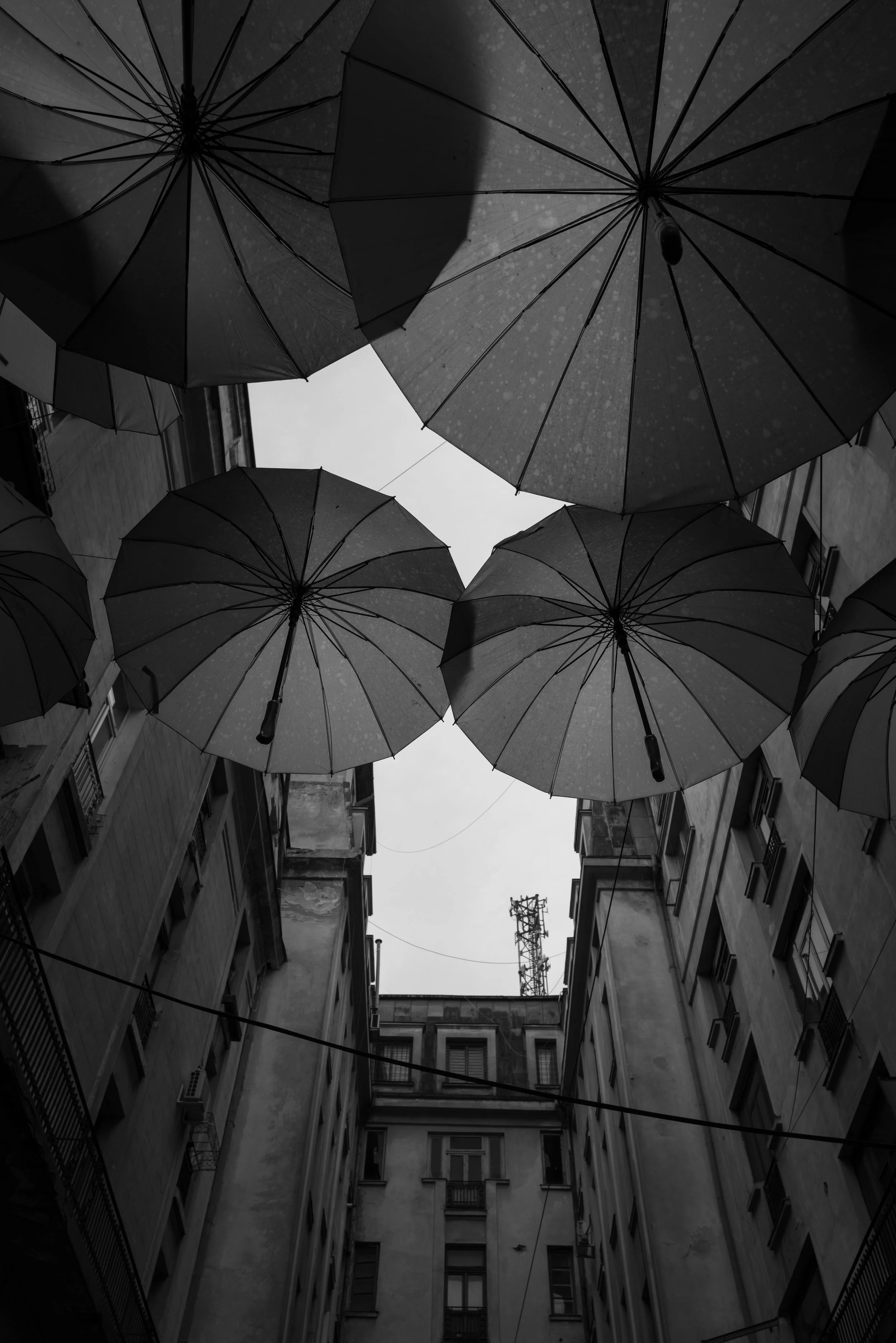 Schwarz-weiß-Foto von Regenschirmen, die über einem Innenhof zwischen mehrstöckigen Gebäuden aufgehängt sind.