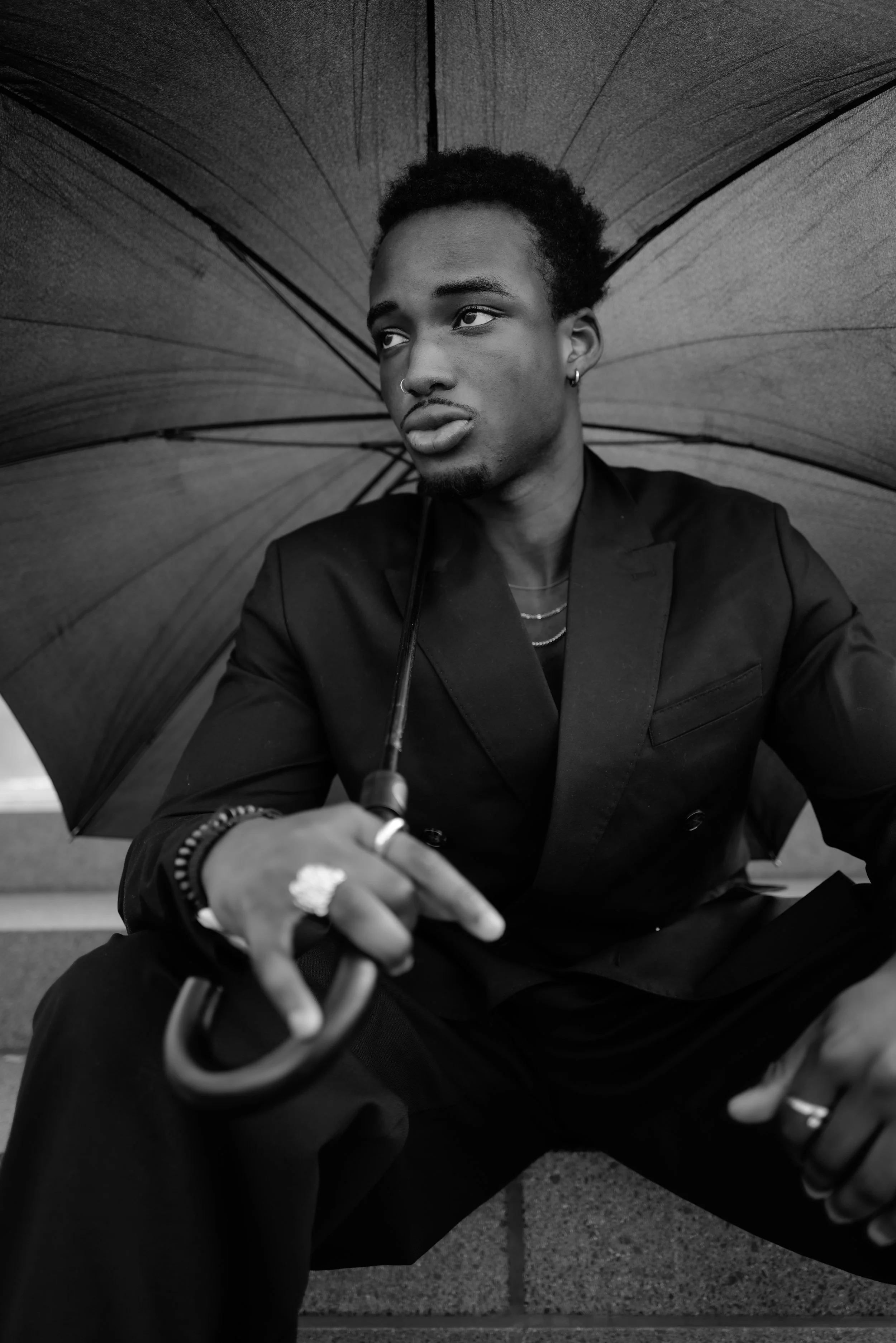 Schwarz-Weiß-Fotografie eines jungen Mannes, der unter einem großen Regenschirm sitzt, mit Piercings und Schmuck, in einem urbanen Umfeld.