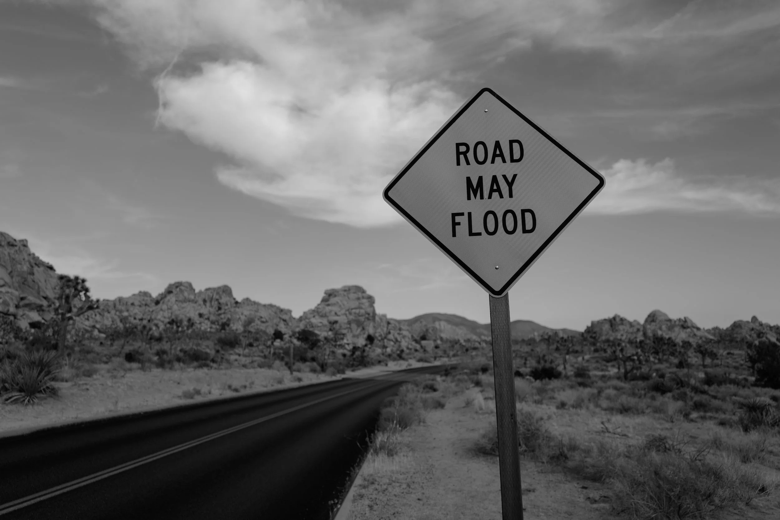 Straßenschild mit der Aufschrift 'Road May Flood' in einer Wüstenlandschaft mit Bergen und Büschen im Hintergrund, Schwarz-Weiß-Bild