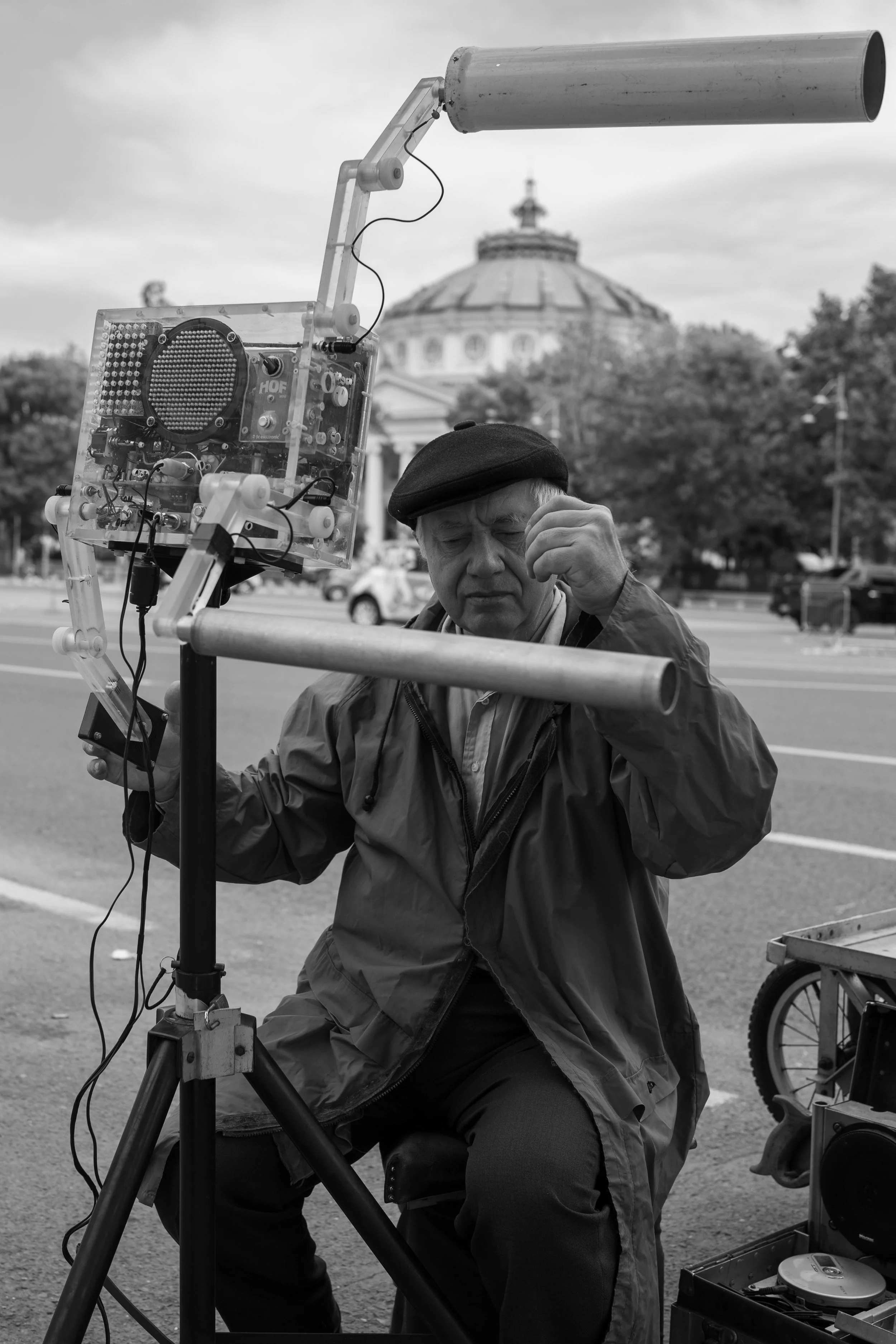 Ein älterer Mann sitzt vor einem ungewöhnlichen Musikinstrument aus Rohren und elektronischen Bauteilen auf einem Stativ. Er trägt eine Mütze und einen Mantel. Im Hintergrund ist ein historisches Gebäude und Bäume zu sehen.