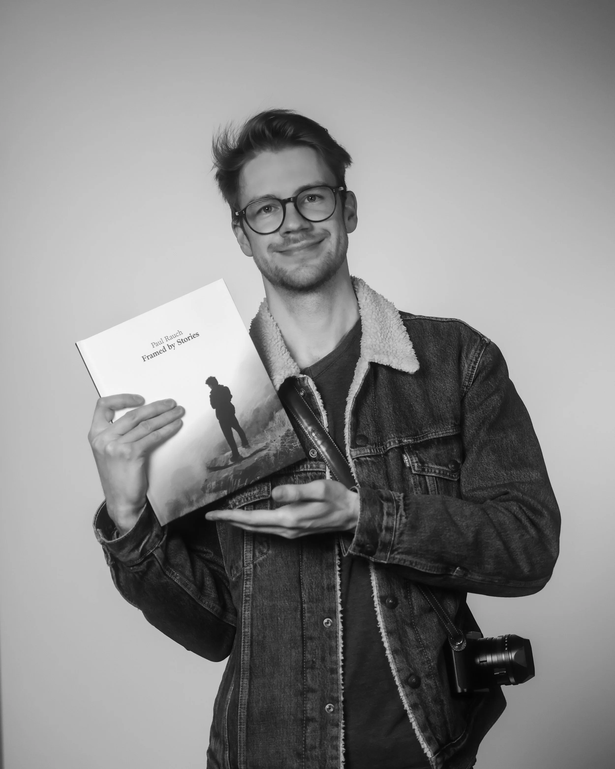 Junge Person mit Brille, trägt eine Jeansjacke, hält ein Buch mit dem Titel 'Framed by Stories' von Paul Rauch, lächelt vor weißem Hintergrund.
