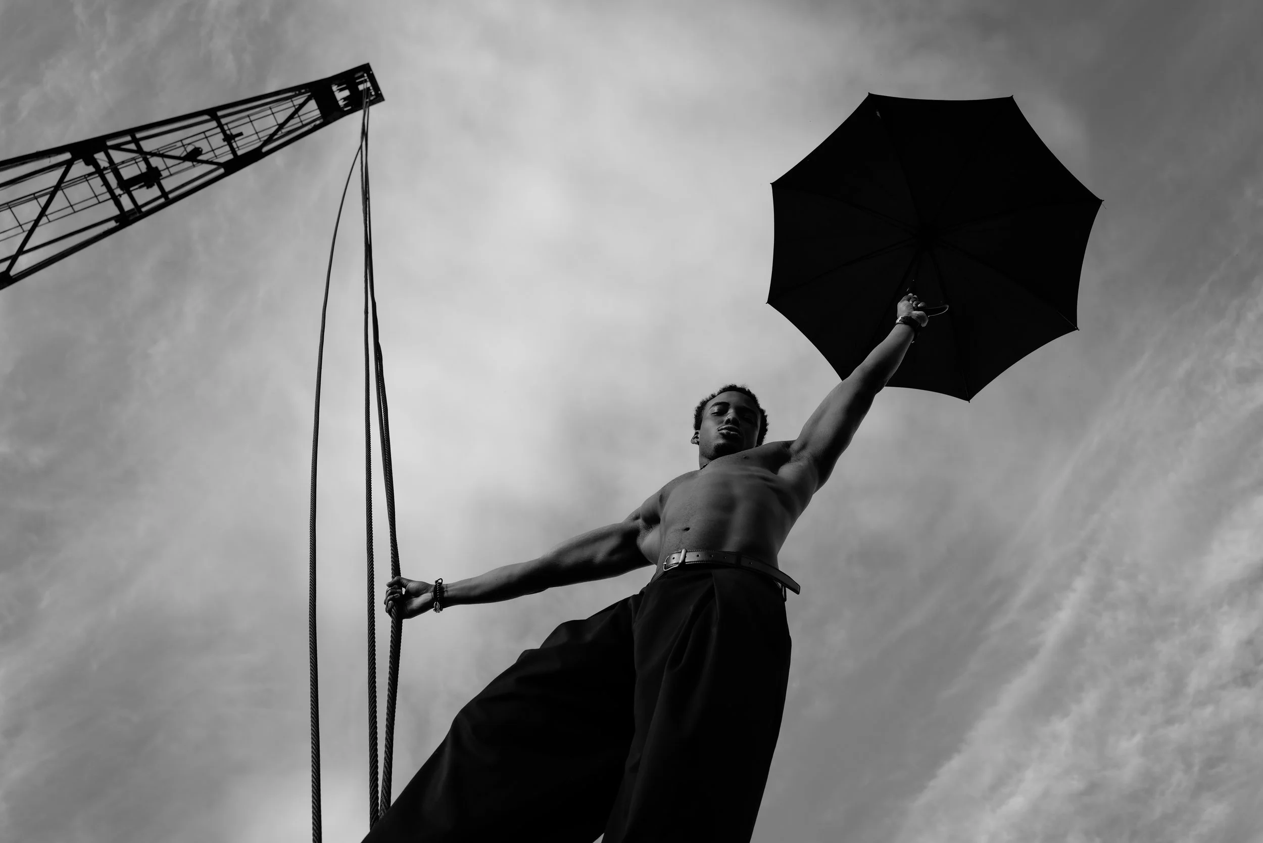 Schwarze Silhouette eines Mannes mit nach oben ausgestrecktem Arm, der ein schwarzes Regenschirm hält, vor einem Himmel mit Wolken, im Hintergrund eine Baukran.
