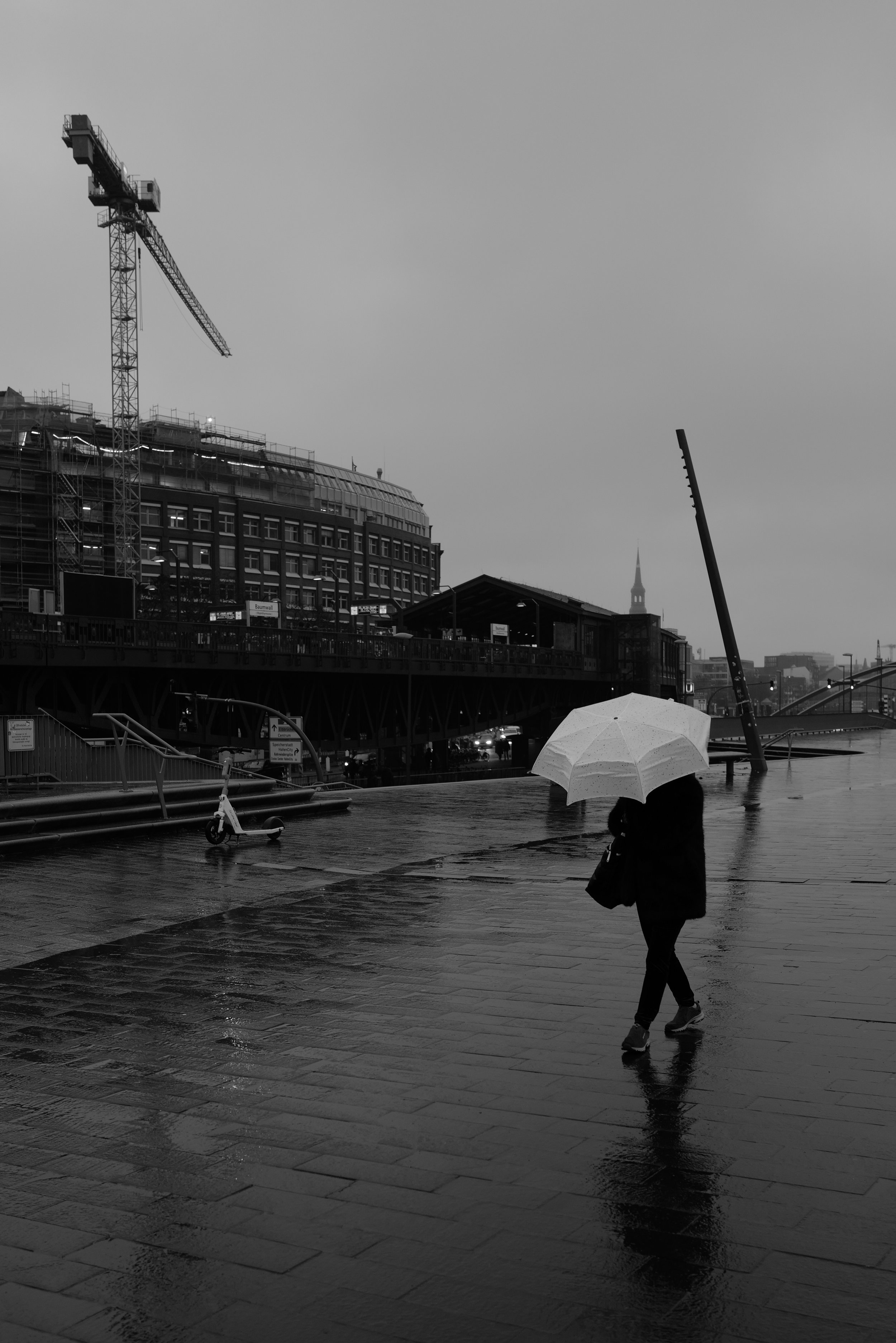 Schwarz-weiß-Foto von einer nassen Stadtlandschaft mit einer Person, die mit einem Regenschirm auf nassem Pflaster geht. Im Hintergrund sind ein unter Bau befindliches Gebäude mit einem Kran und eine städtische Architektur zu sehen.